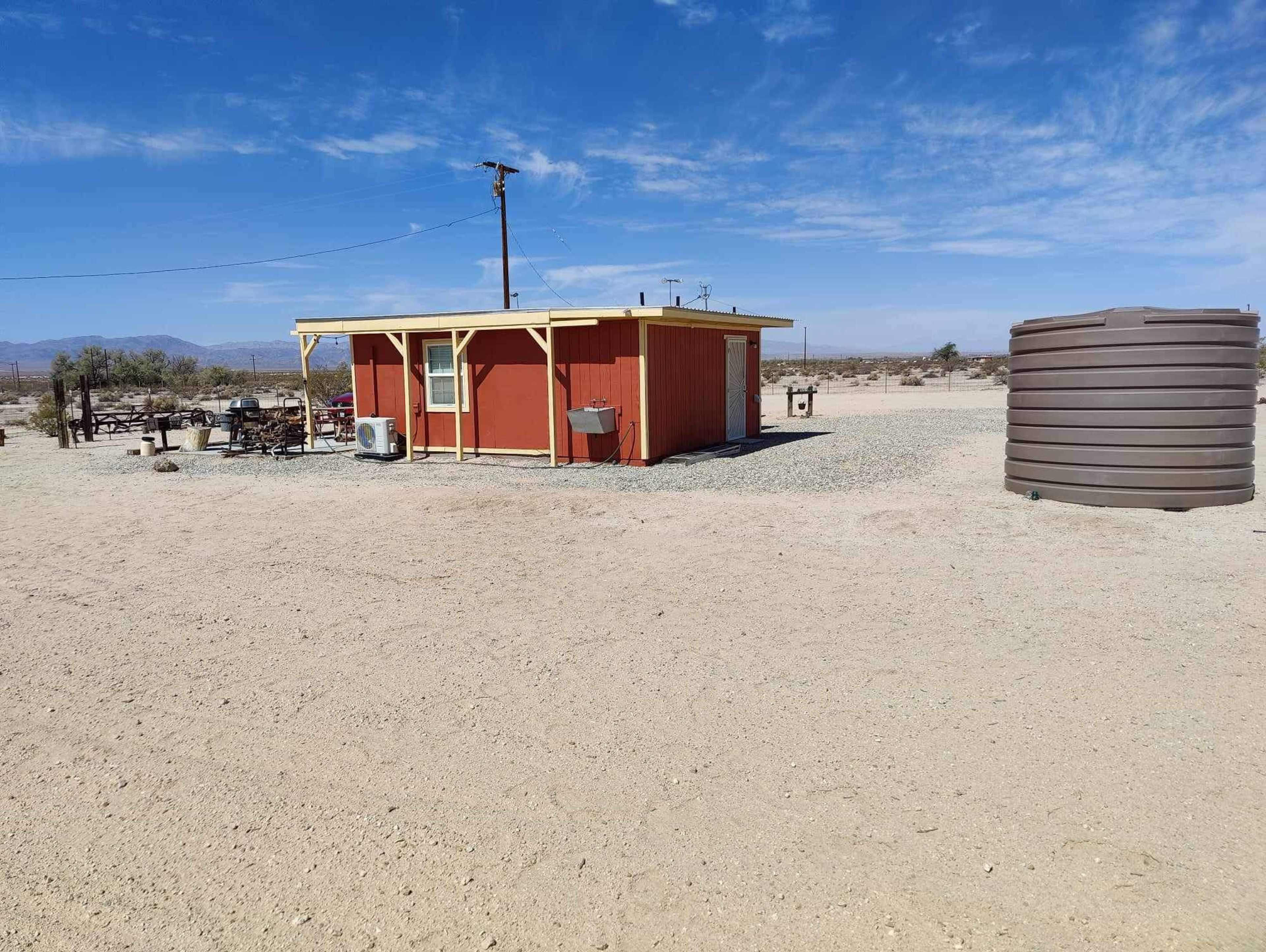 A small red building is situated in a sandy, open area beside a large water tank.