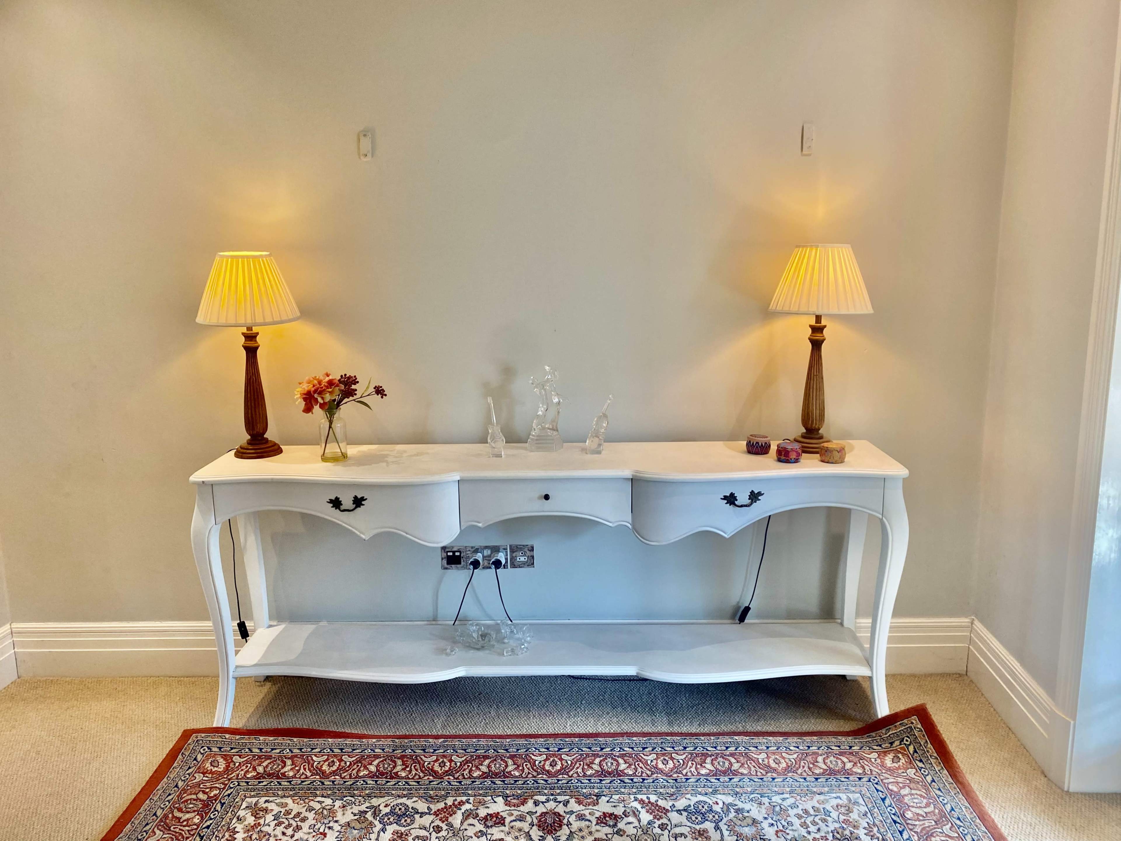 A white console table with two lamps and decorative items sits against a light-colored wall.