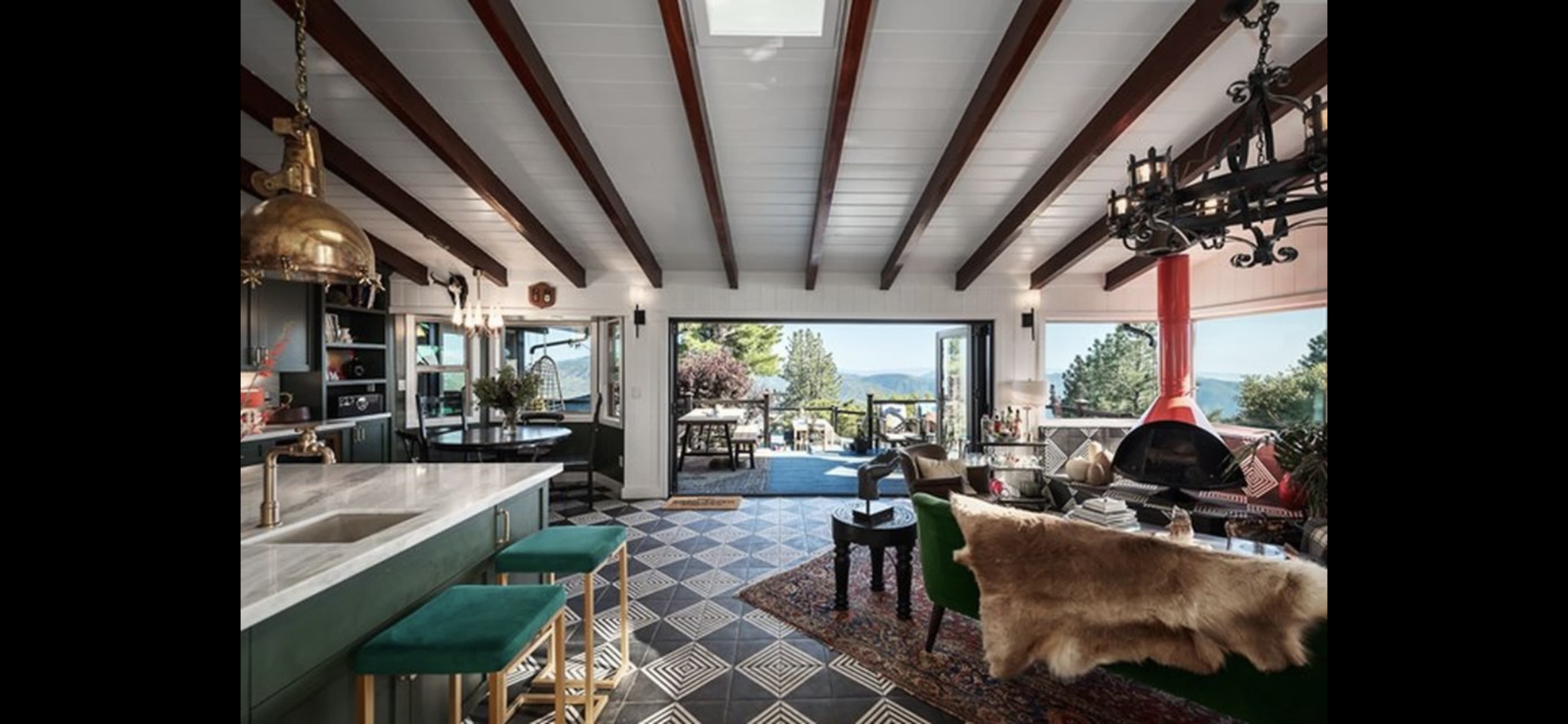A spacious living area with wooden beam ceilings, a kitchen island, and large windows revealing a scenic view of the outdoors.