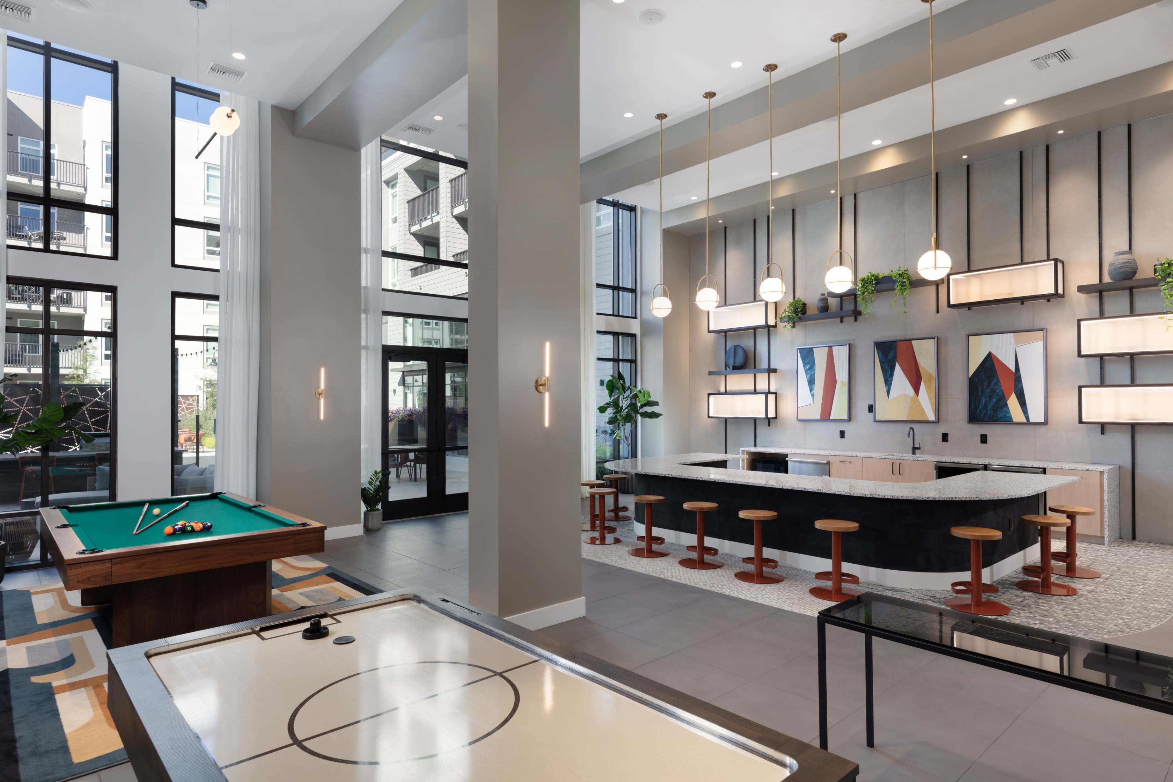 The image shows a modern communal lounge area featuring a pool table, air hockey table, and a bar counter with stools, surrounded by large windows and decorative artwork.