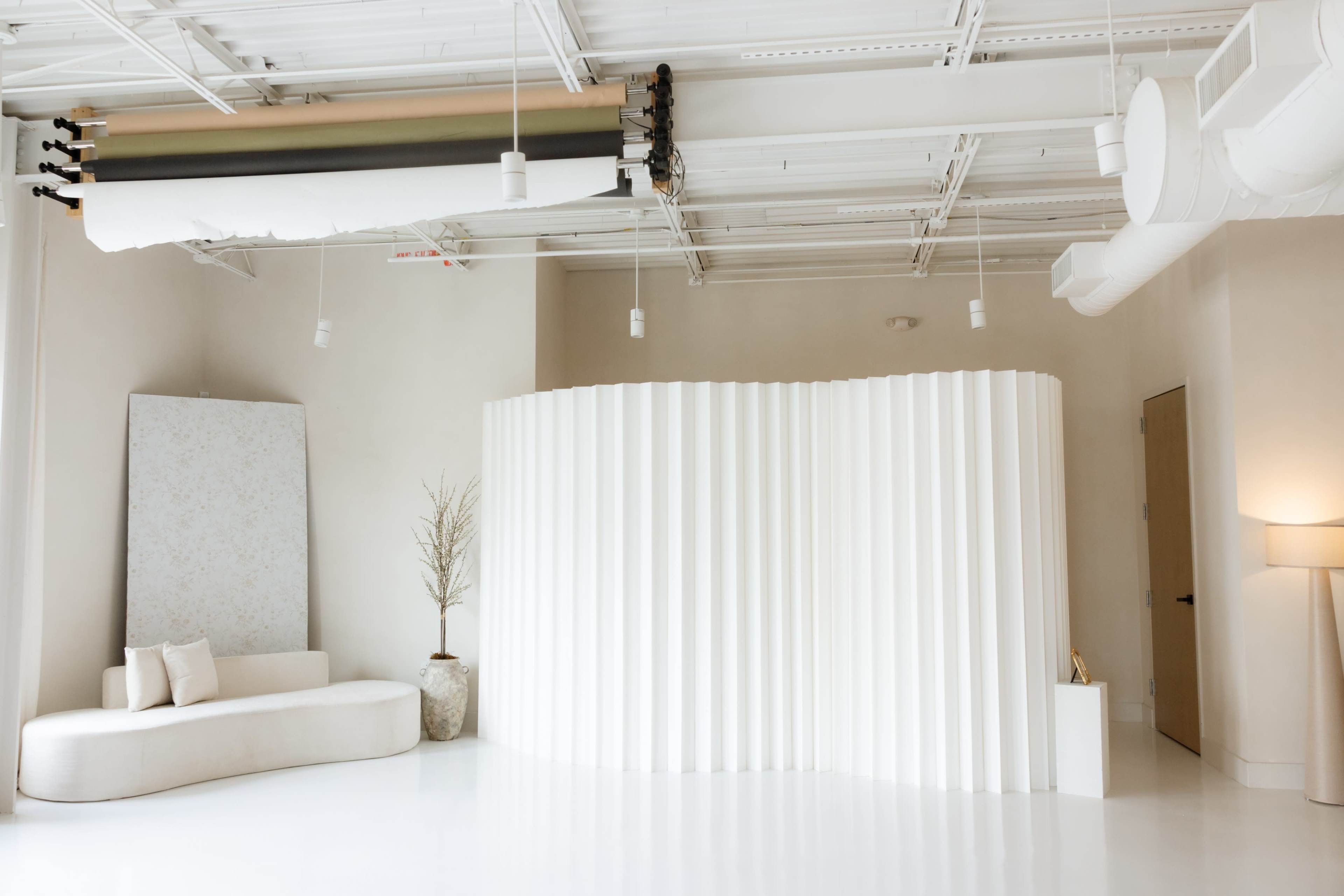 The image shows a modern, minimalist interior space featuring a curved white partition, a light couch, a decorative plant, and soft-colored walls.