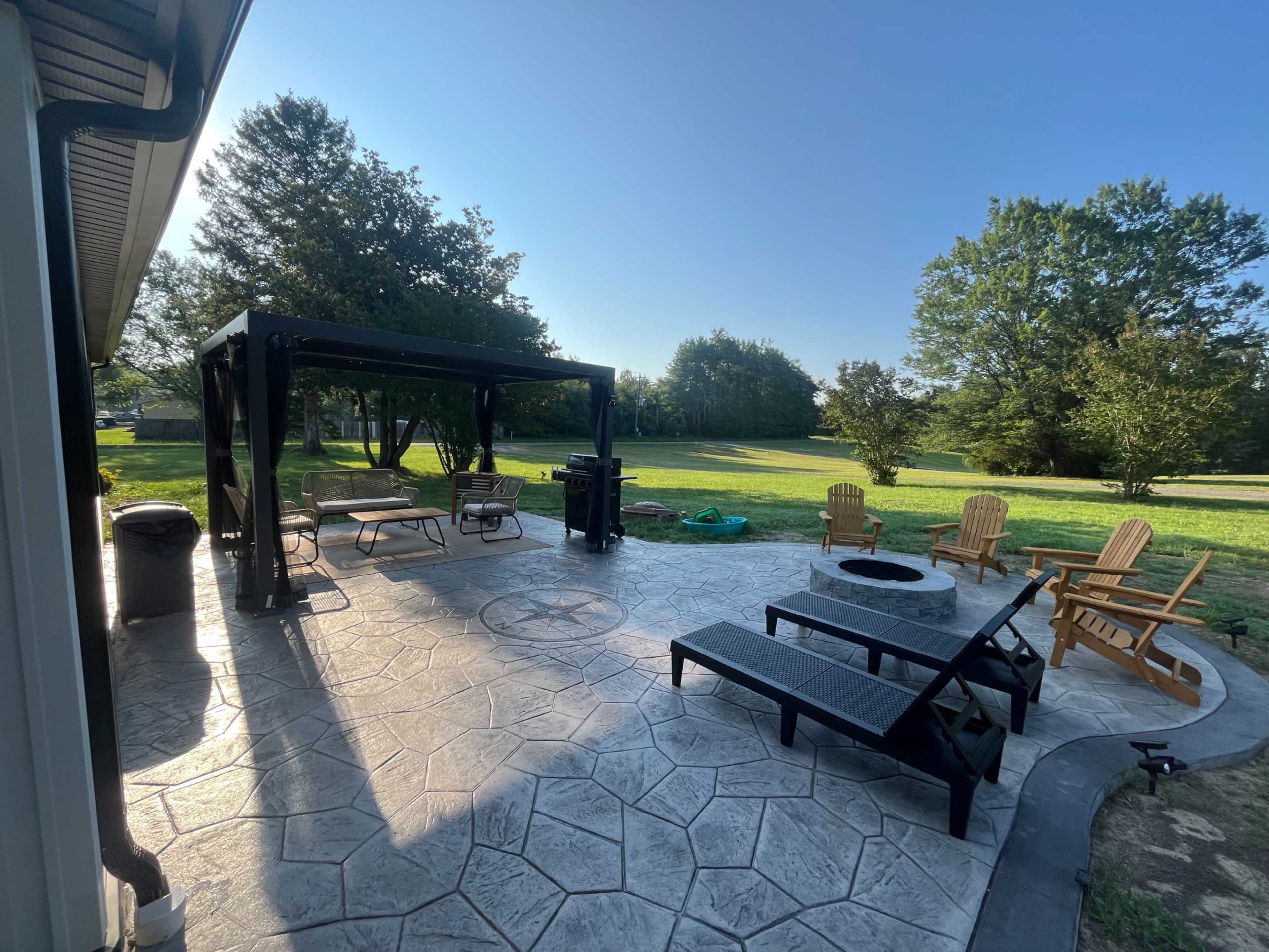 The image shows a backyard patio with a gazebo, outdoor seating, and a fire pit, surrounded by a grassy lawn and trees.