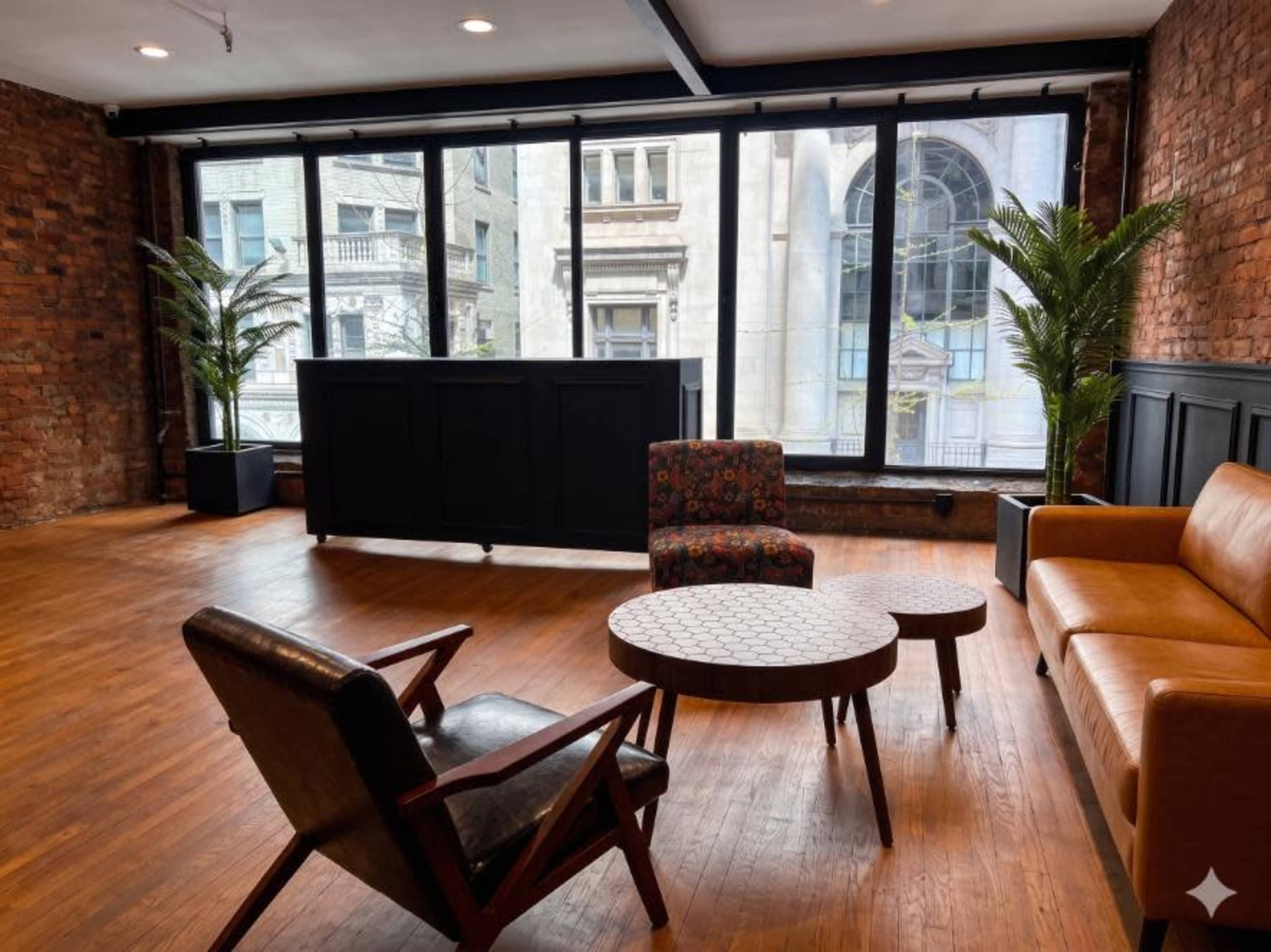 The image shows a snug interior space with large windows, wooden flooring, a small circular coffee table, and a mix of modern seating arrangements including a brown sofa and an armchair.