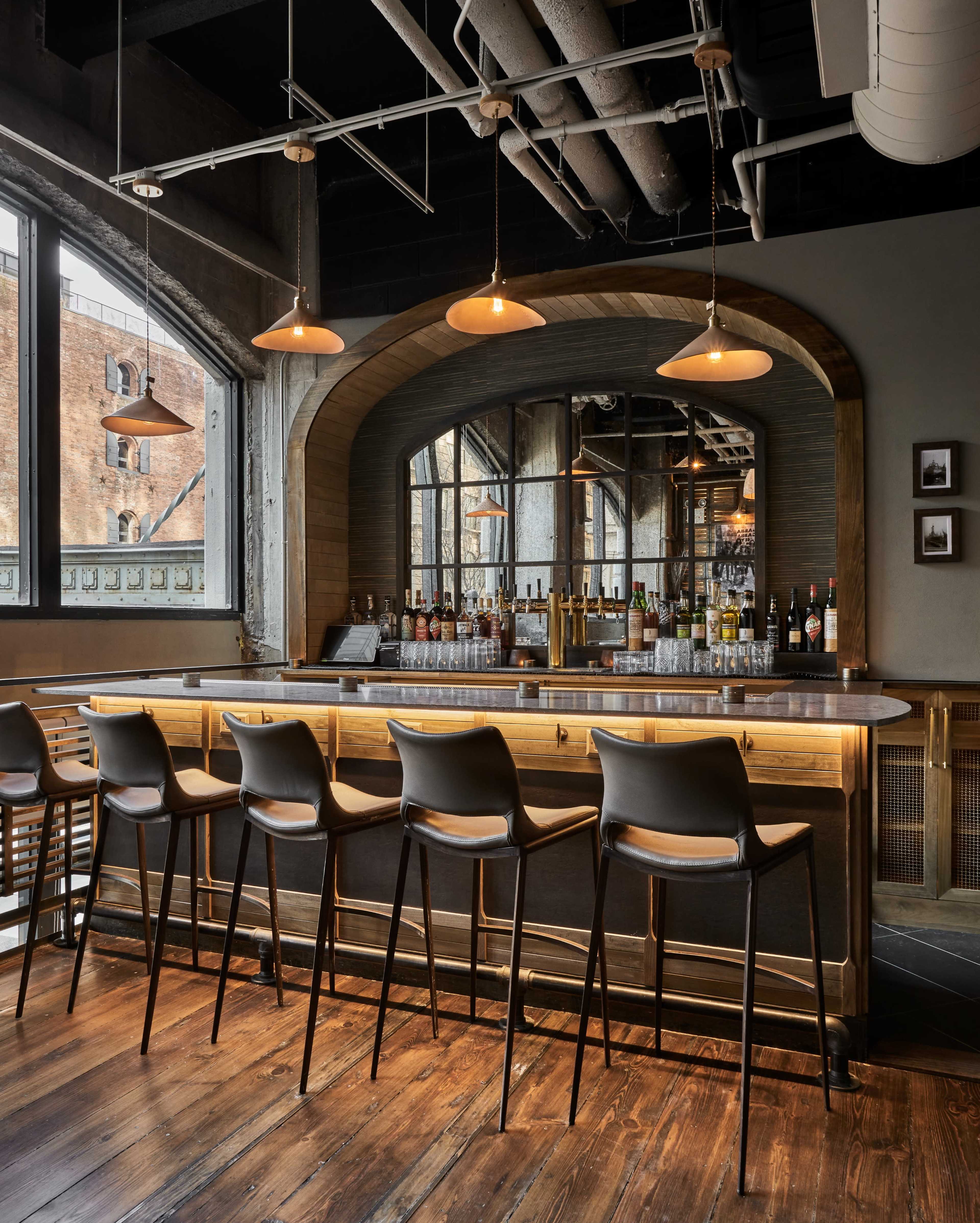 A modern bar with a wooden counter and high stools, set against a backdrop of large windows and industrial decor.