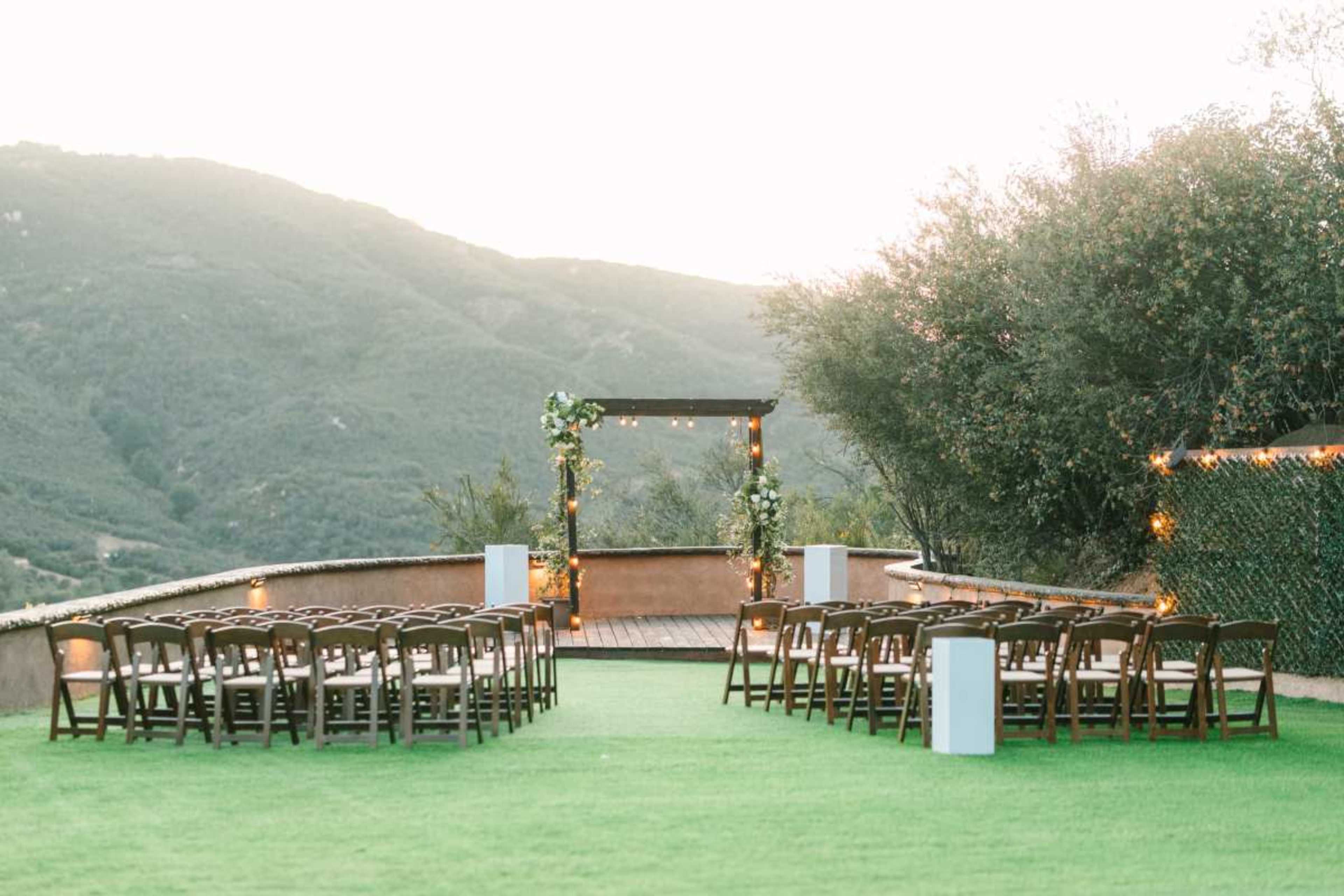 An outdoor wedding ceremony setup features rows of wooden chairs facing a decorated arch, set against a mountainous backdrop.