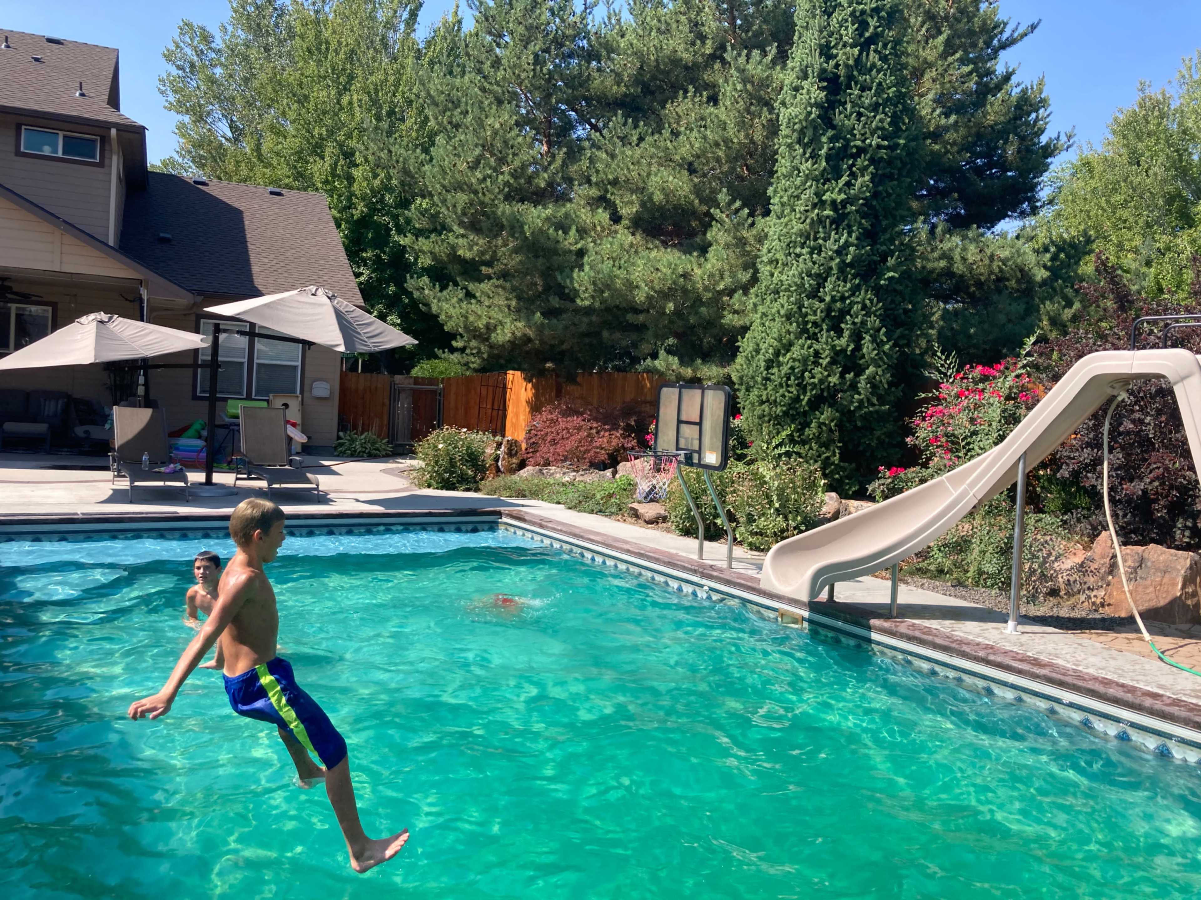 A boy jumps into a swimming pool with a slide in a residential backyard surrounded by trees and landscaping.