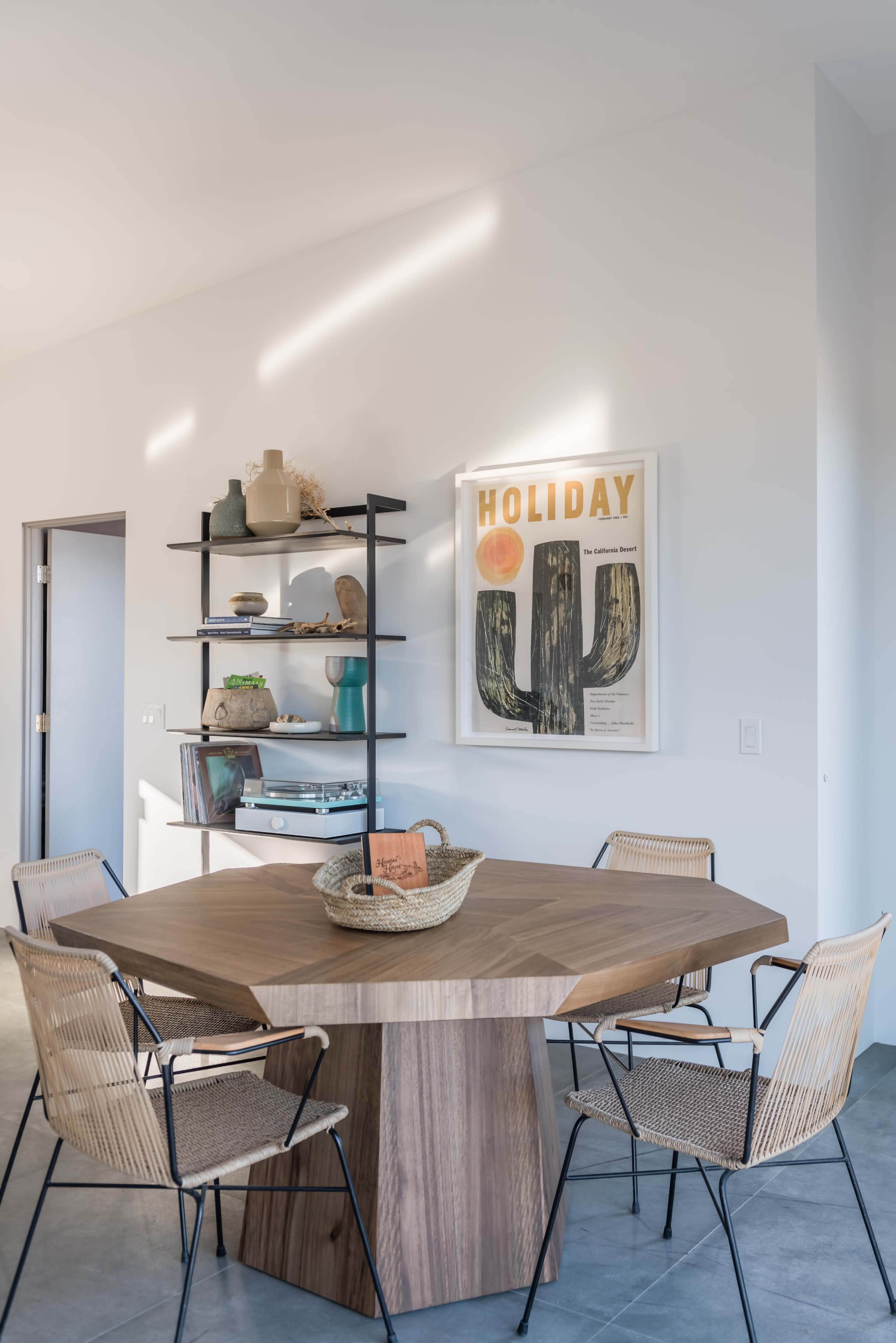 A modern dining area features a hexagonal wooden table surrounded by wireframe chairs, with a wall shelf displaying decorative items and a framed artwork above.