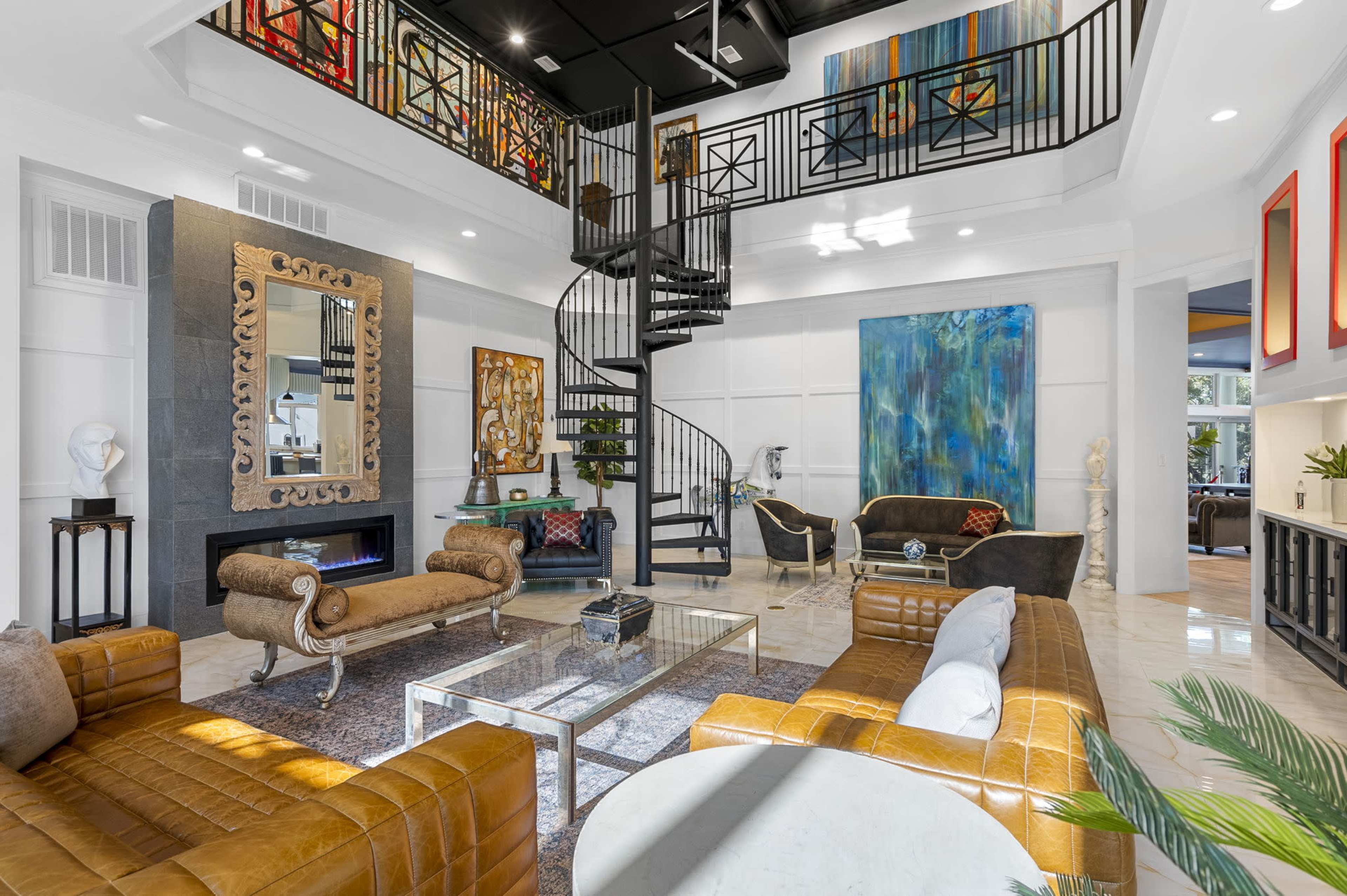 A spacious and modern living room featuring a spiral staircase, a large mirror, and various artworks on the walls, with elegant furnishings arranged around a coffee table.