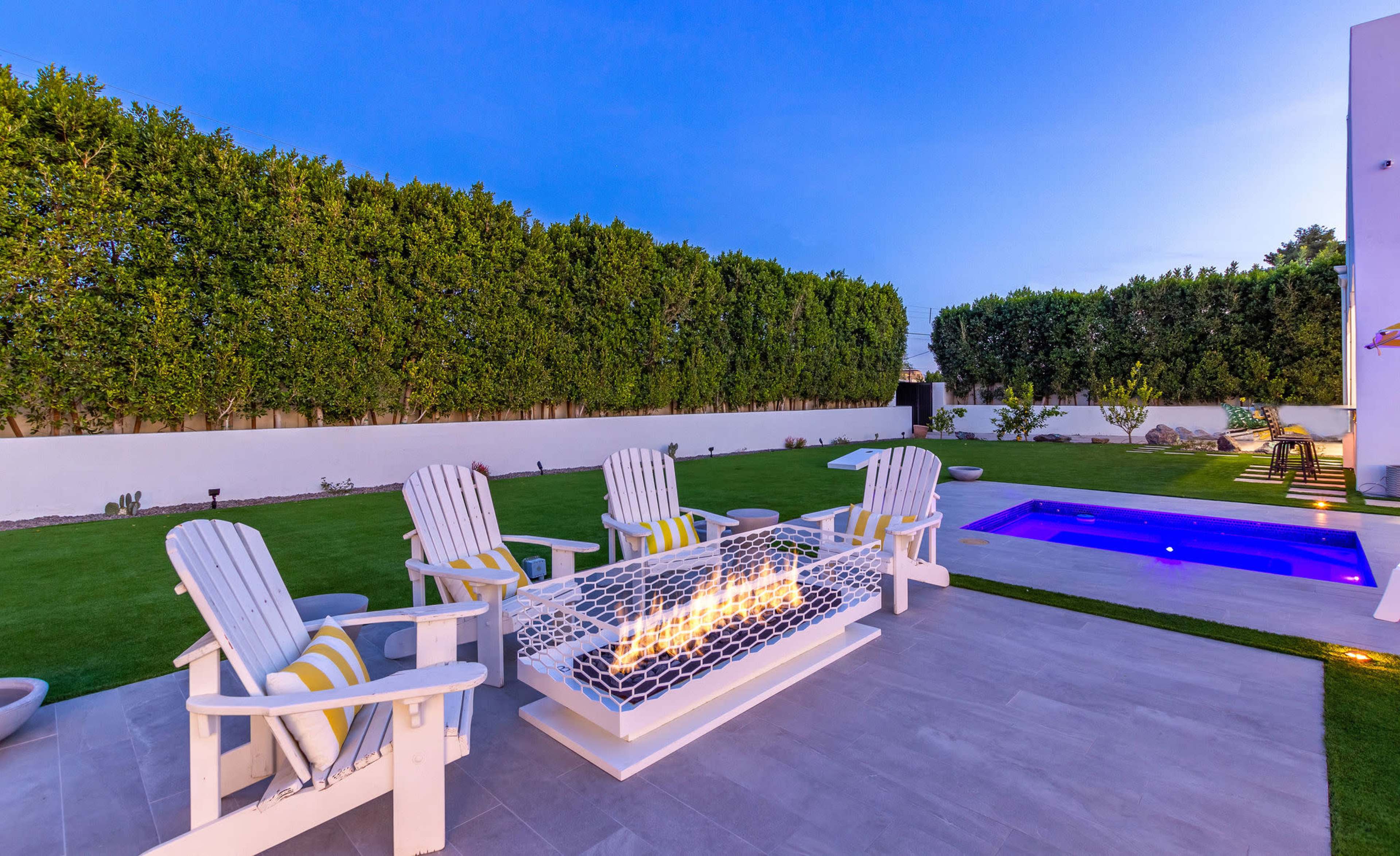 A backyard scene with five white Adirondack chairs arranged around a fire pit, a small swimming pool in the corner, and neatly trimmed hedges along a white wall.