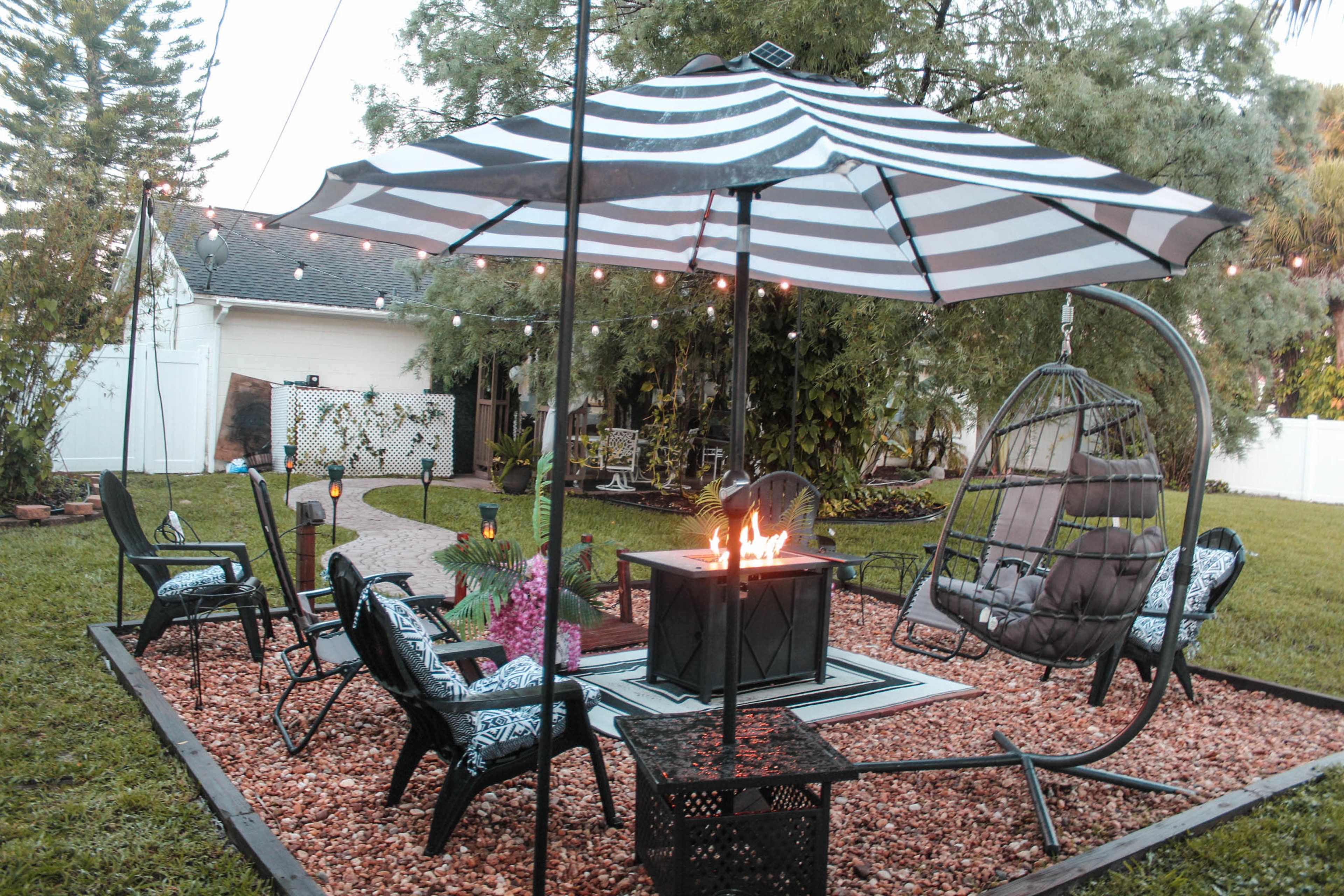 A patio area features a striped umbrella over a fire pit, surrounded by black chairs and a swinging lounge chair, with landscaping and string lights in the background.