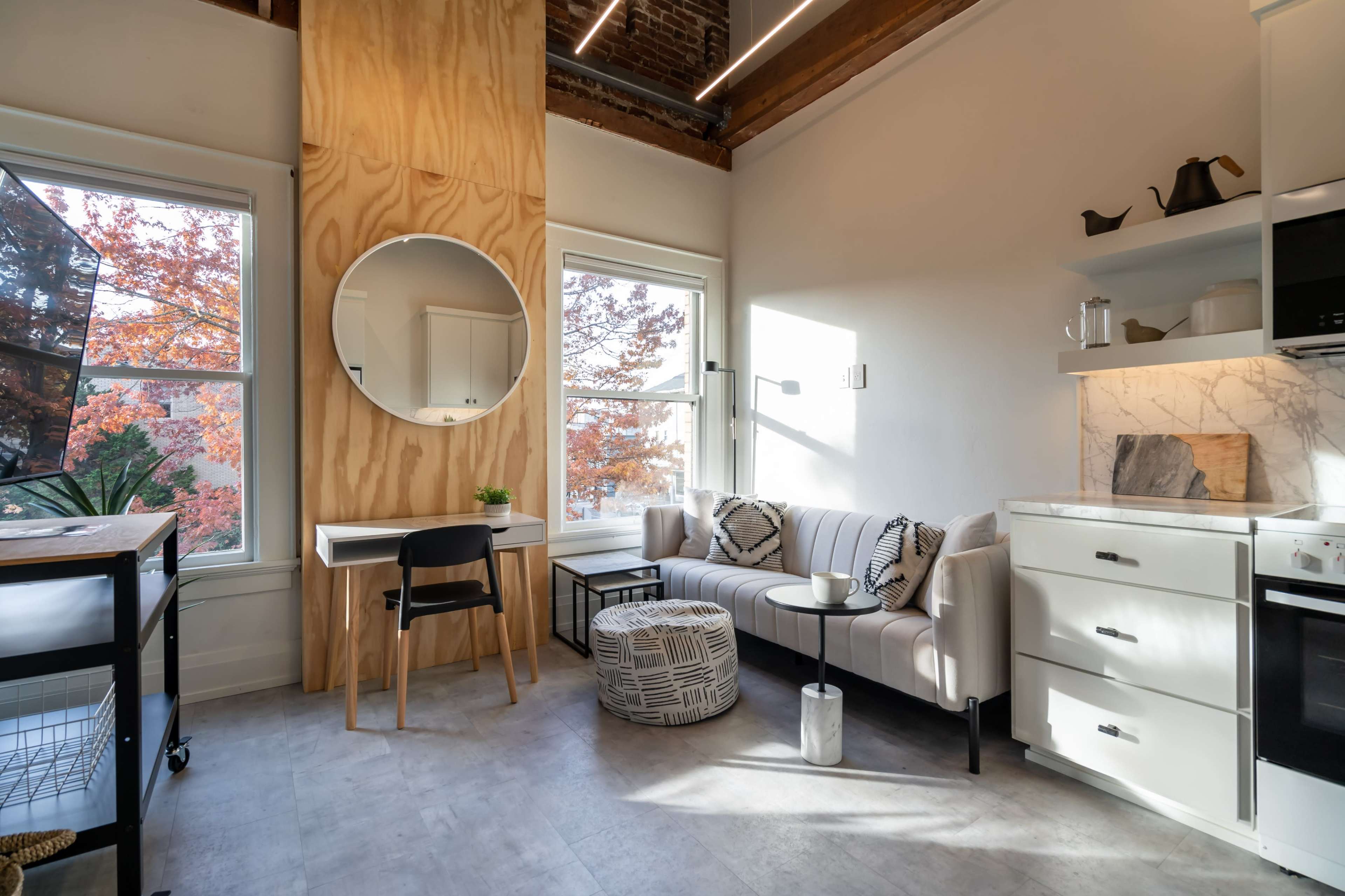 A small, modern living space with a sofa, a round coffee table, a desk with a mirror, and large windows revealing autumn foliage outside.