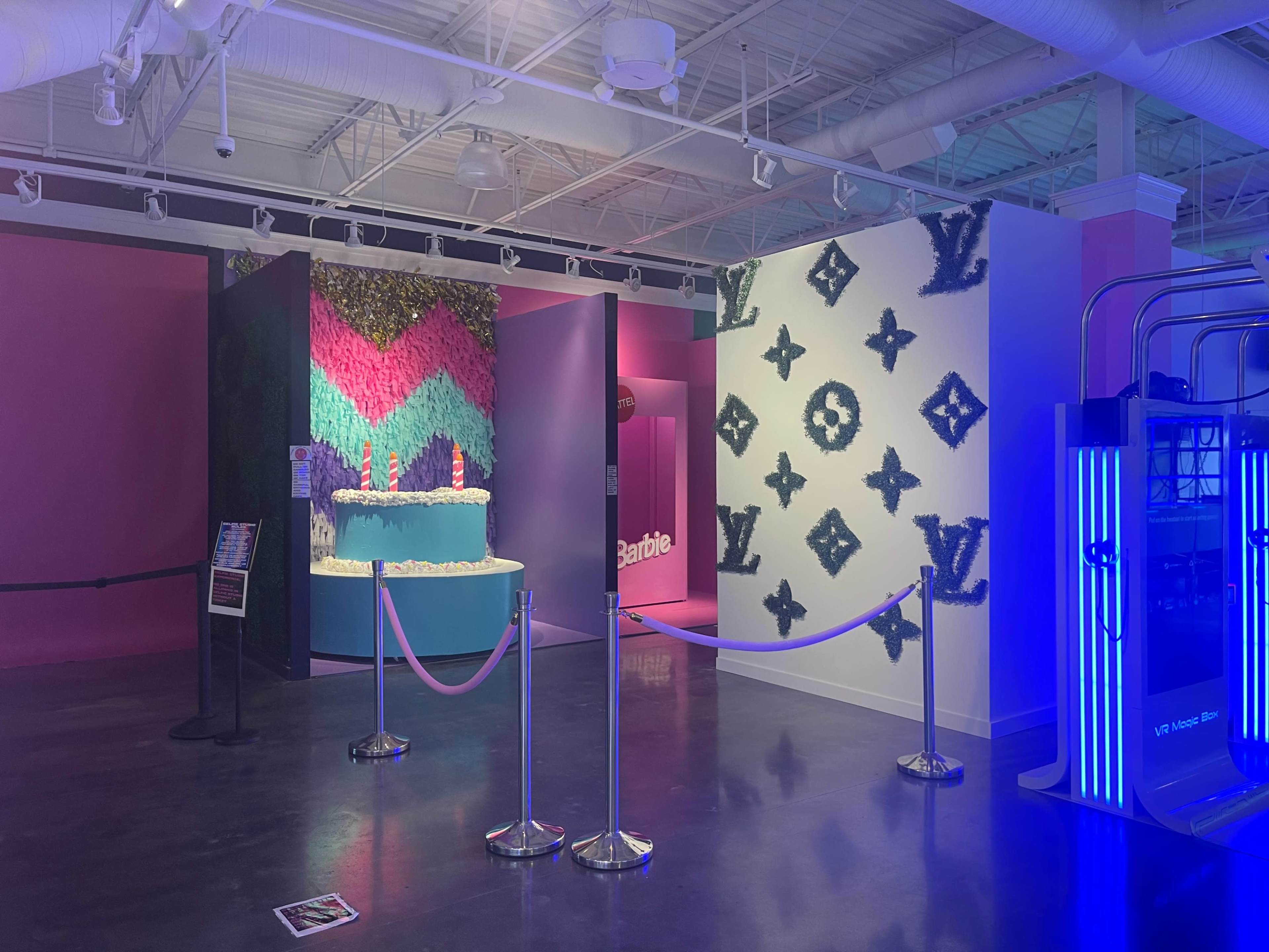 An interior space featuring vibrant walls with a pink and green zigzag design, a blue display with the "Barbie" logo, and a white wall adorned with a Louis Vuitton pattern.