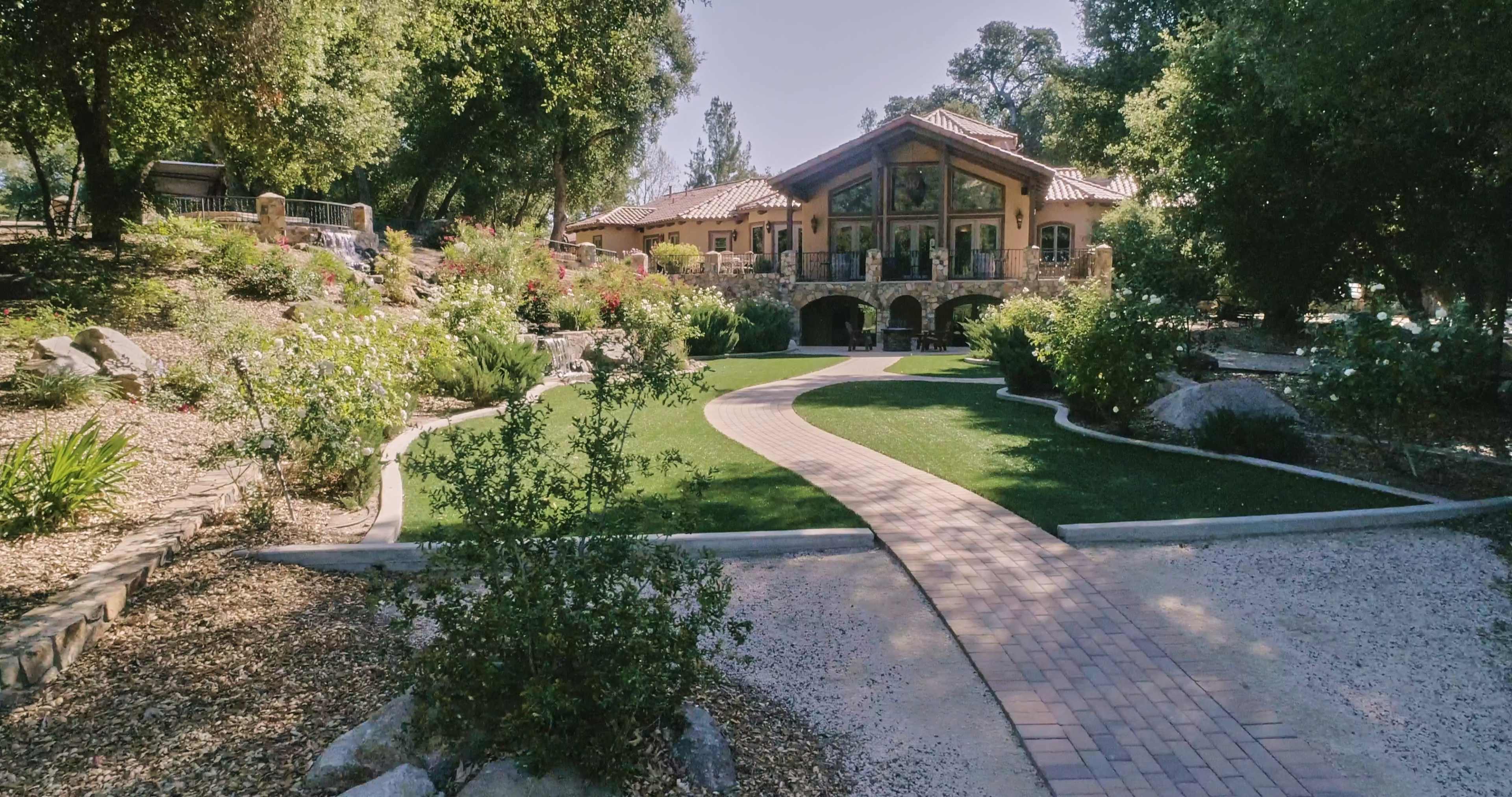 A paved pathway curves through a landscaped garden leading to a large house with a tiled roof.