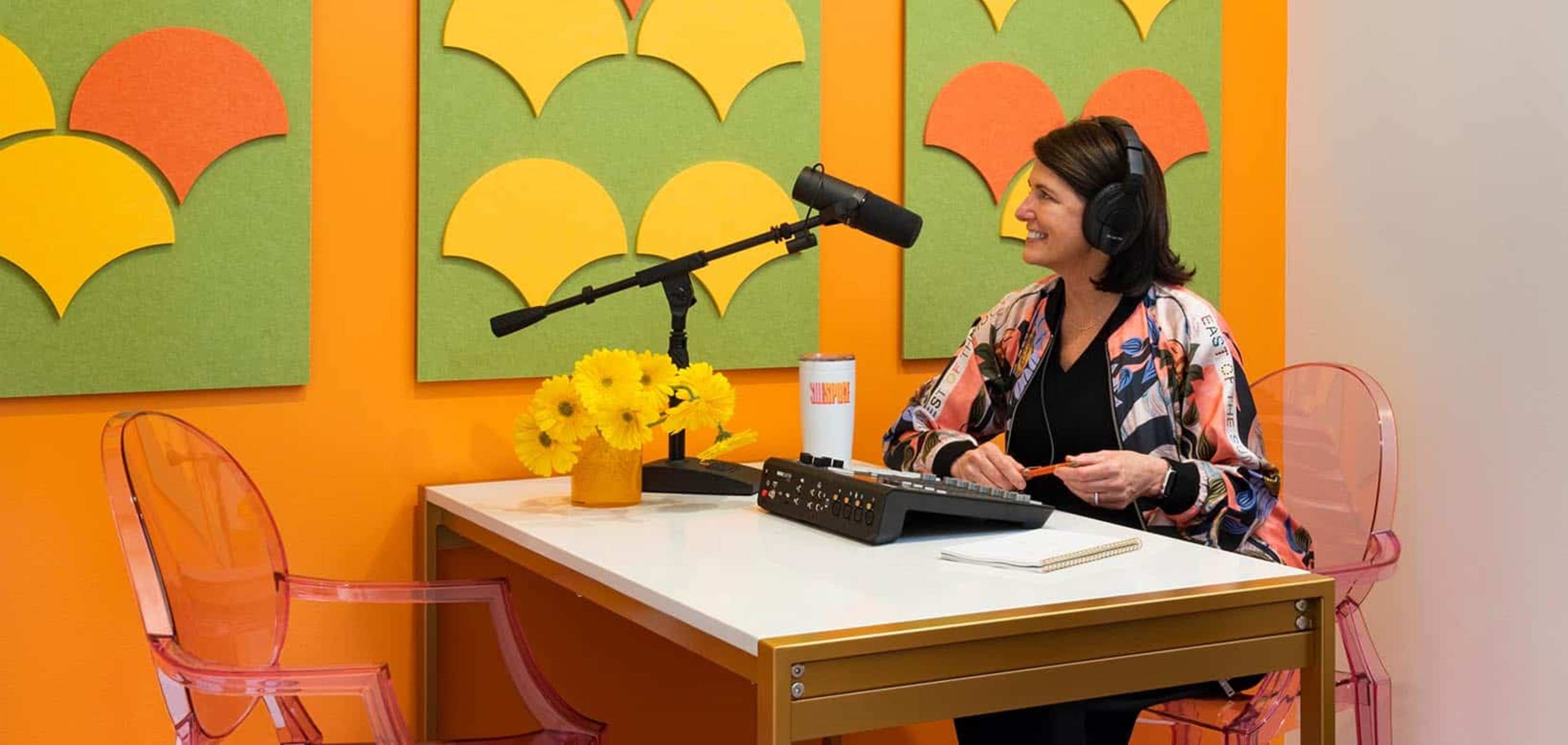 A woman sits at a desk in a brightly colored room, wearing headphones and smiling, with a microphone in front of her and a vase of yellow flowers on the table.