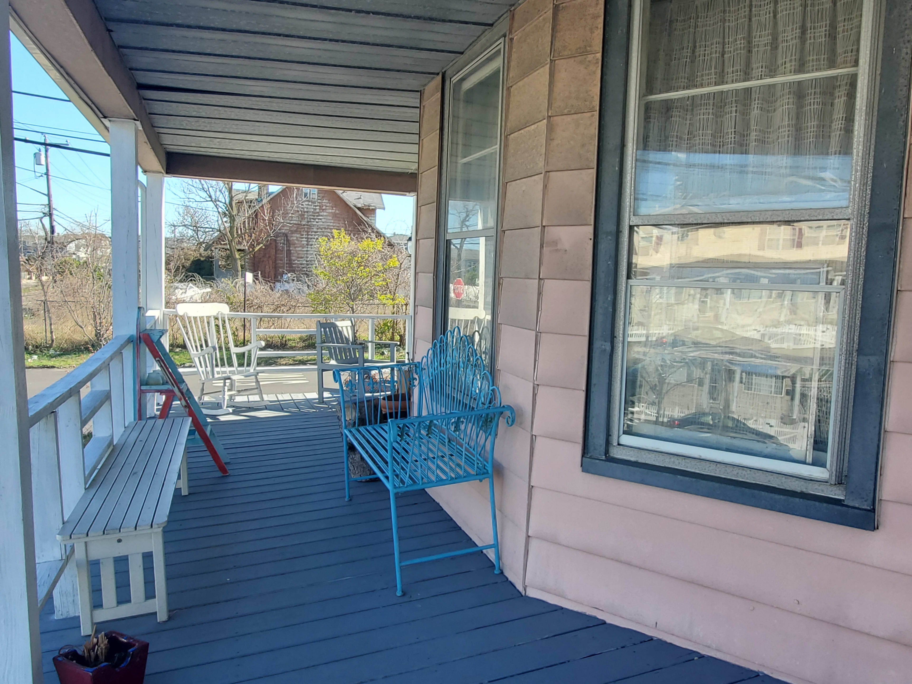 A porch with a blue metal bench, a white wooden bench, and a view of a yard with trees and a house in the background.
