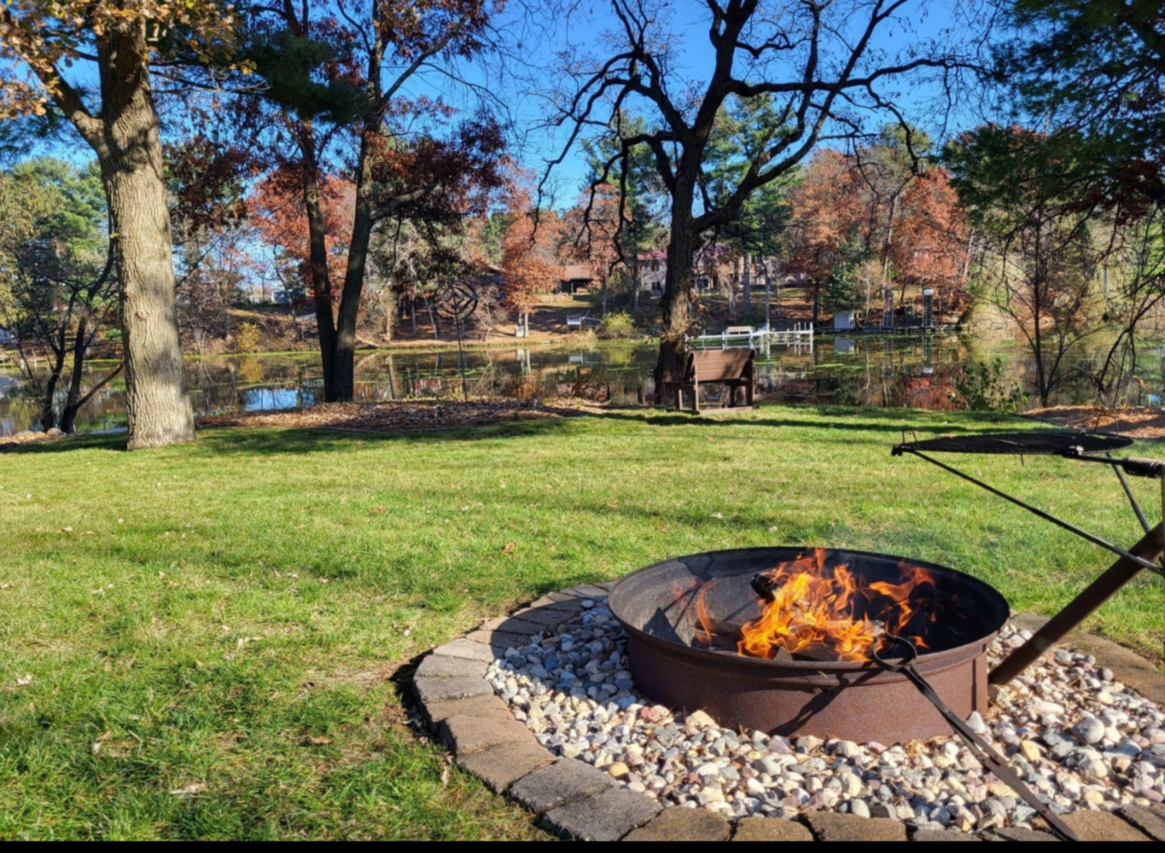 A fire pit with flames is surrounded by a stone border on a grassy area overlooking a pond and trees in autumn foliage.