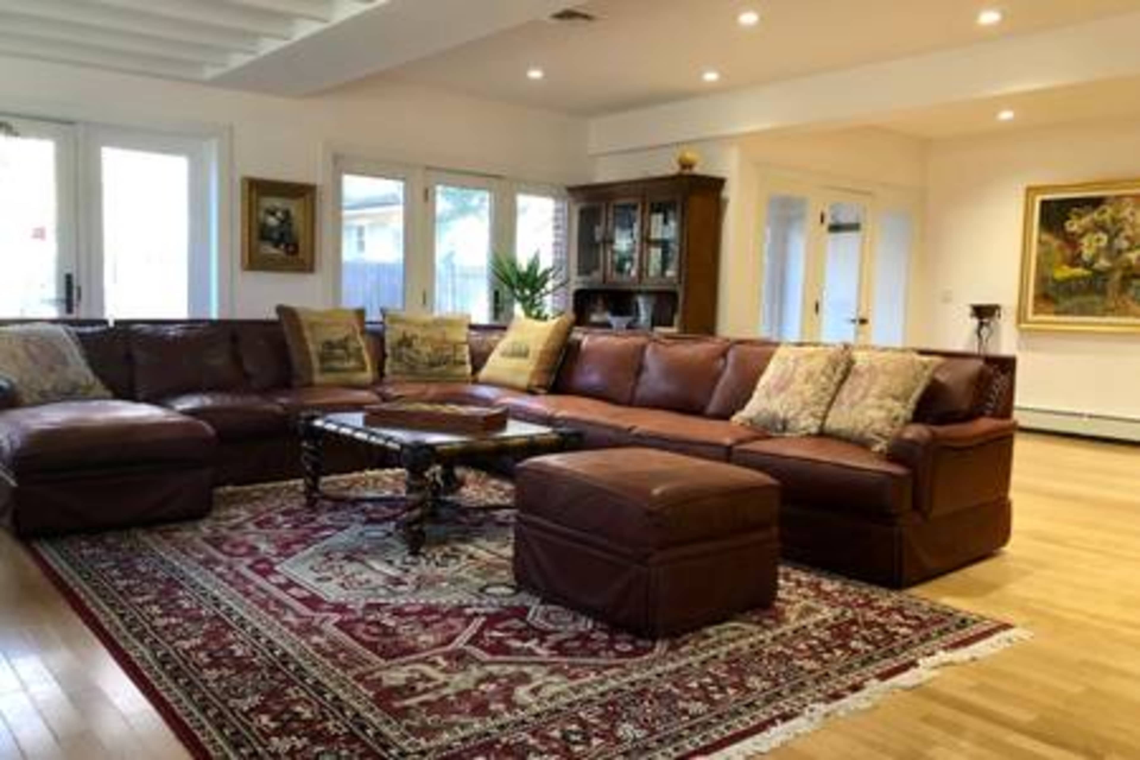 A spacious living room features a large brown leather sectional couch, a decorative coffee table, and an ornate area rug under bright lighting.