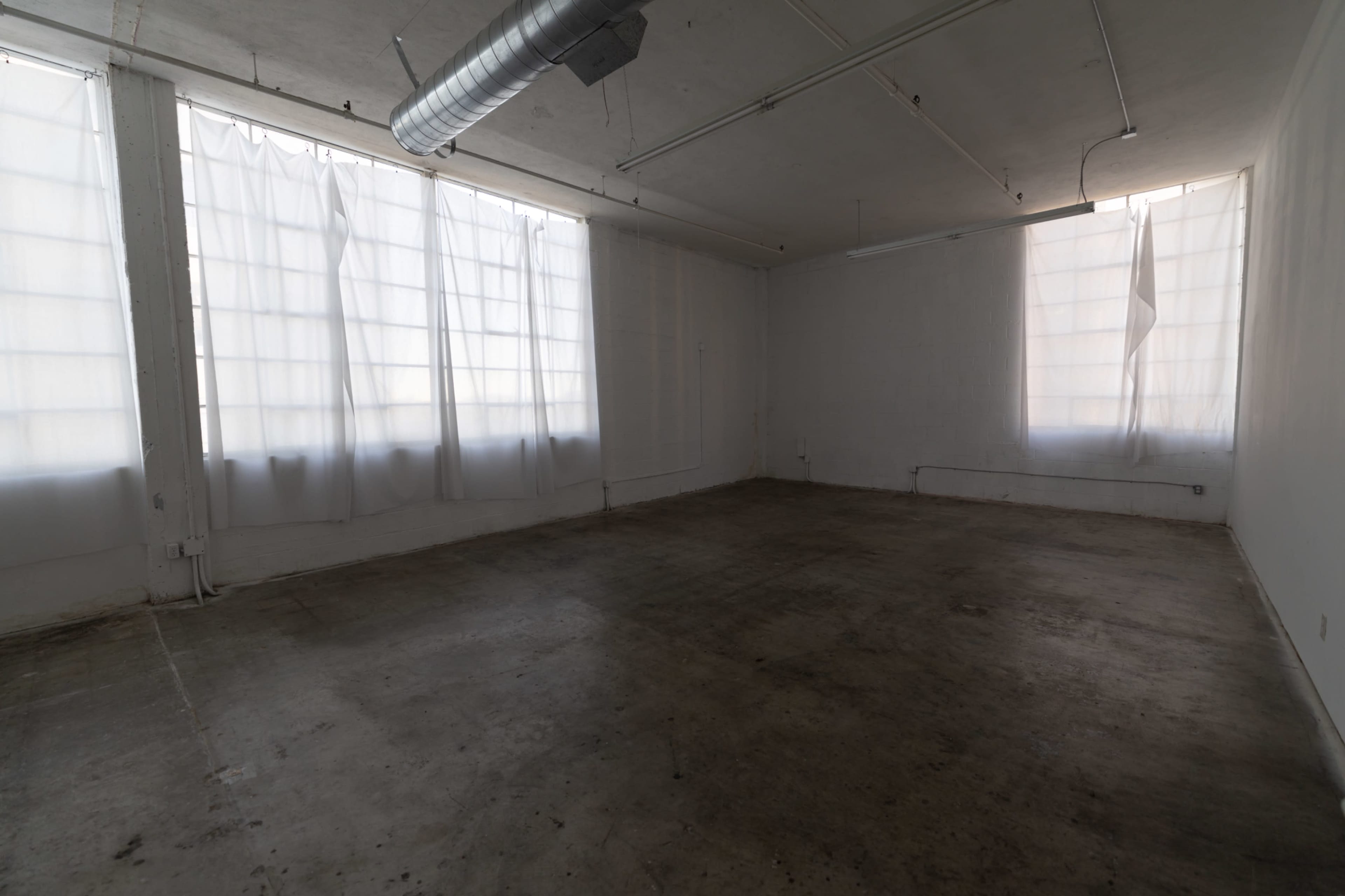 The image shows an empty room with concrete flooring and large, partially draped windows that allow diffuse light to enter.