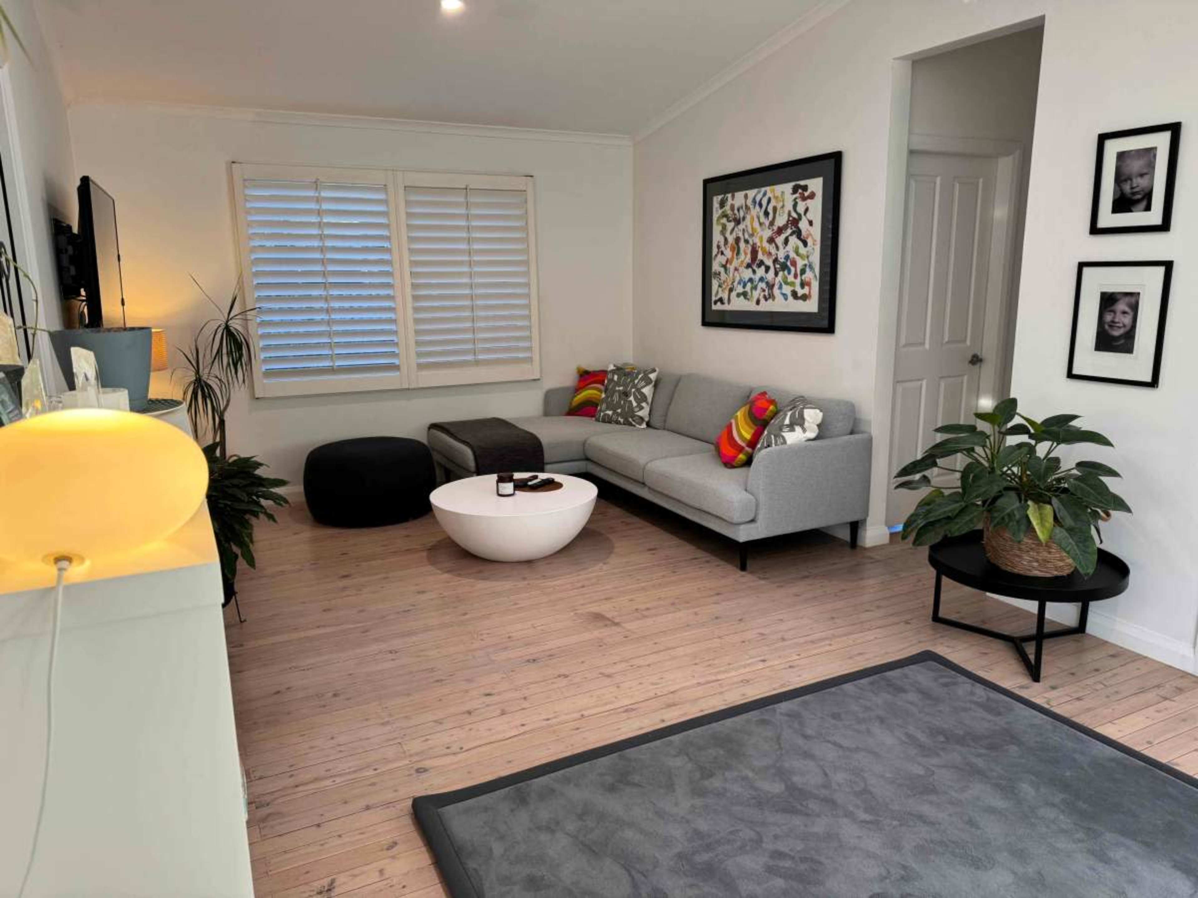 The image shows a modern living room featuring a gray sofa, a round white coffee table, a black ottoman, and decorative plants, with framed artwork on the walls.