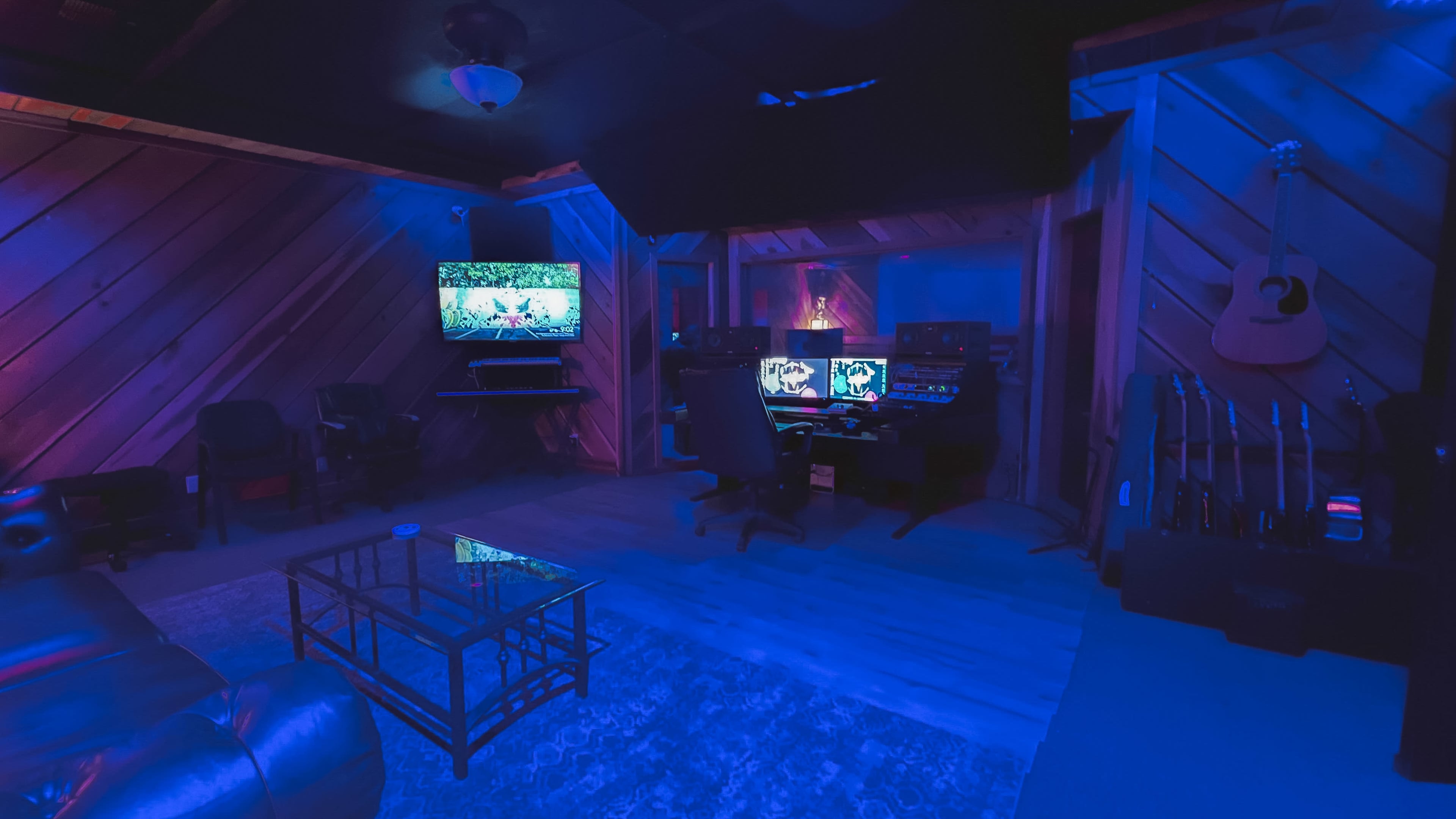 A dimly lit music studio with a computer workstation, a television mounted on the wall, and several musical instruments displayed in the background.