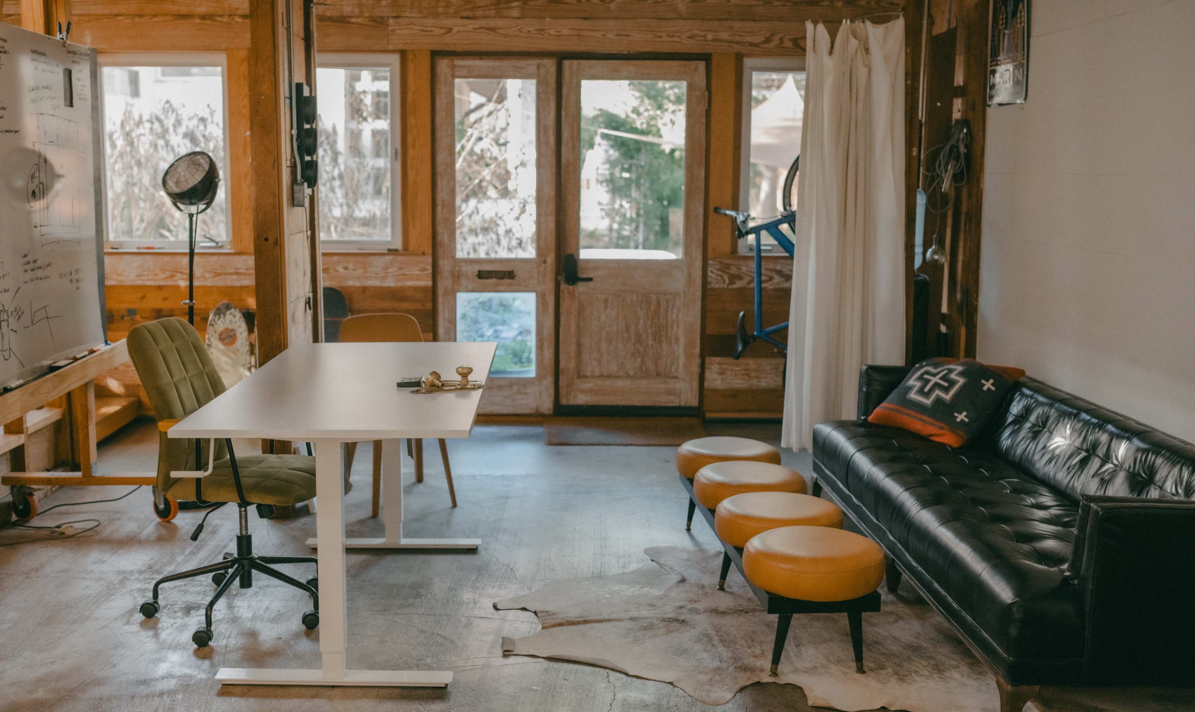 The image shows a modern, minimalist workspace with a wooden interior, featuring a table, chairs, and a black leather sofa.