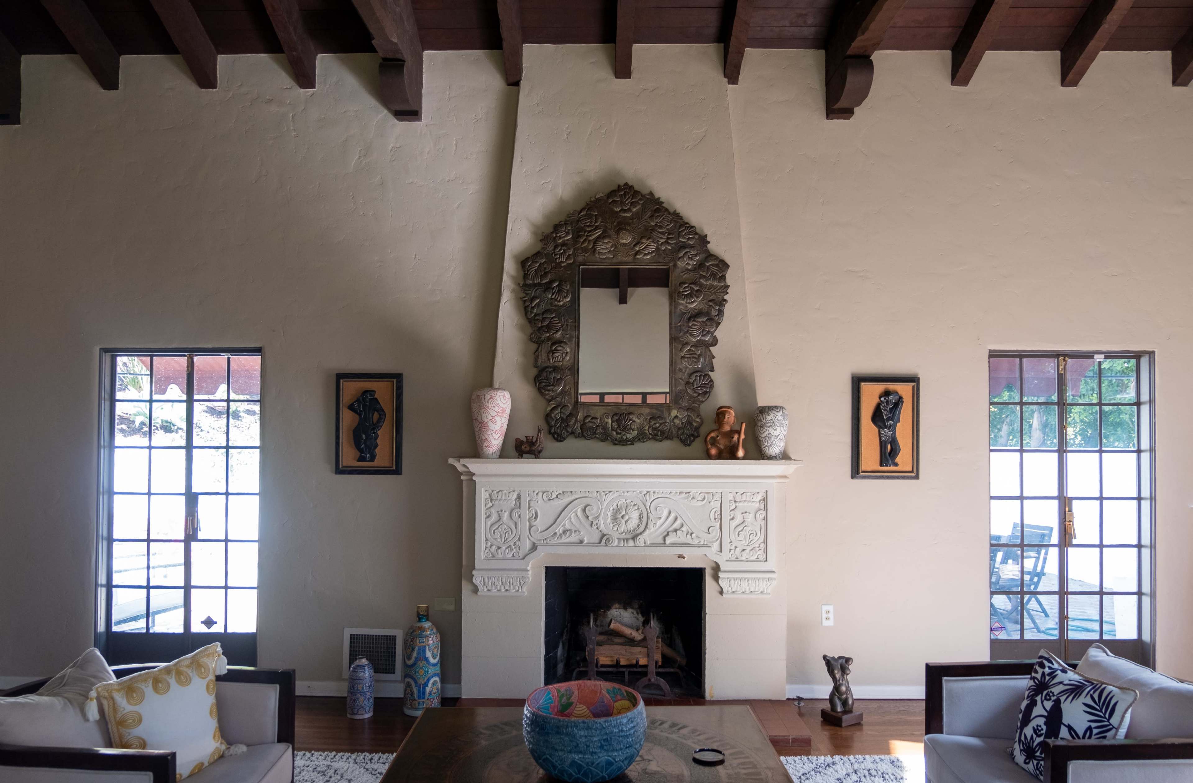 A living room features a decorative fireplace with an ornate mirror above it, flanked by framed artworks and windows on either side.
