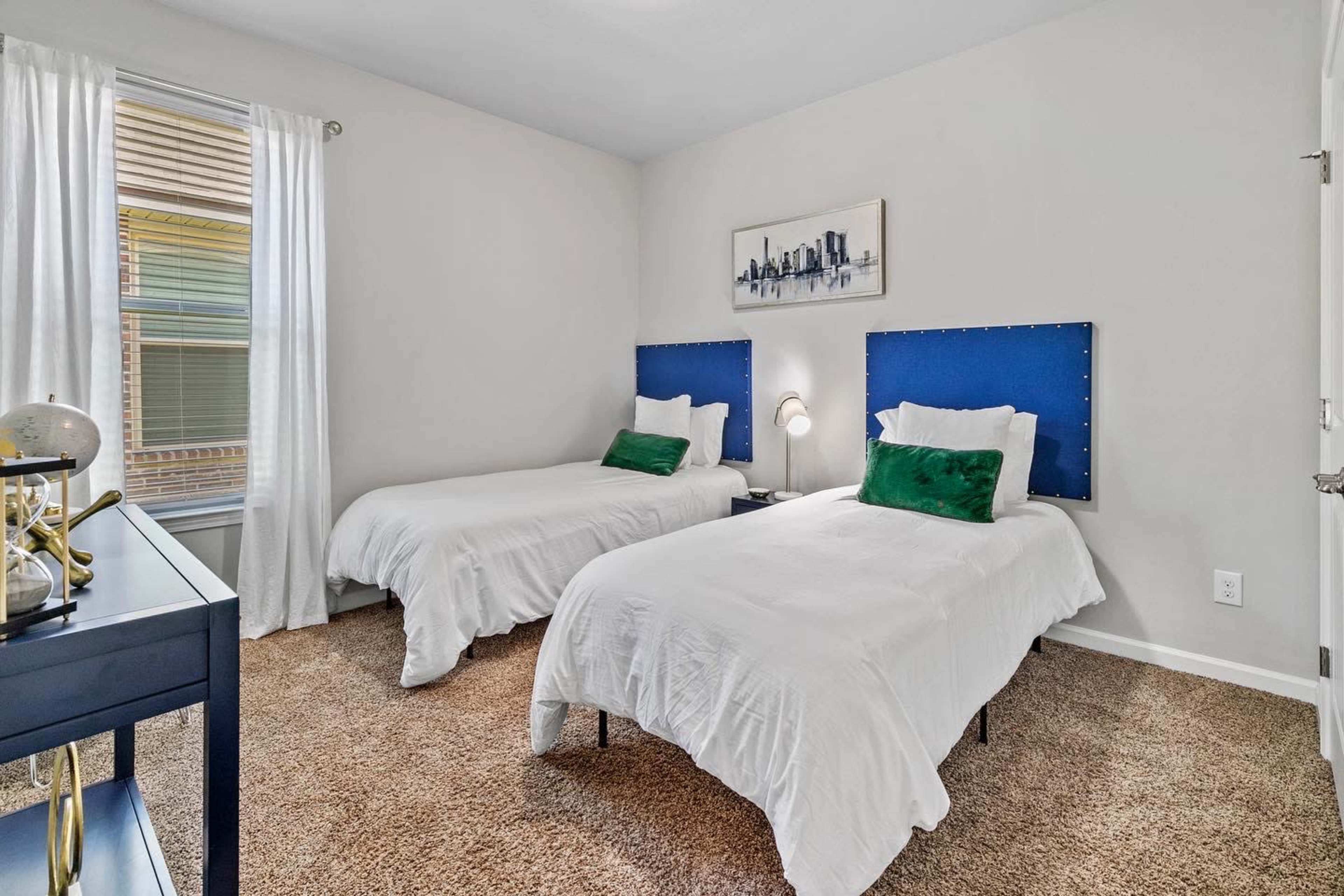 A room with two twin beds, each covered with white bedspreads, and a small table beside them, featuring decorative items and a window with white curtains.
