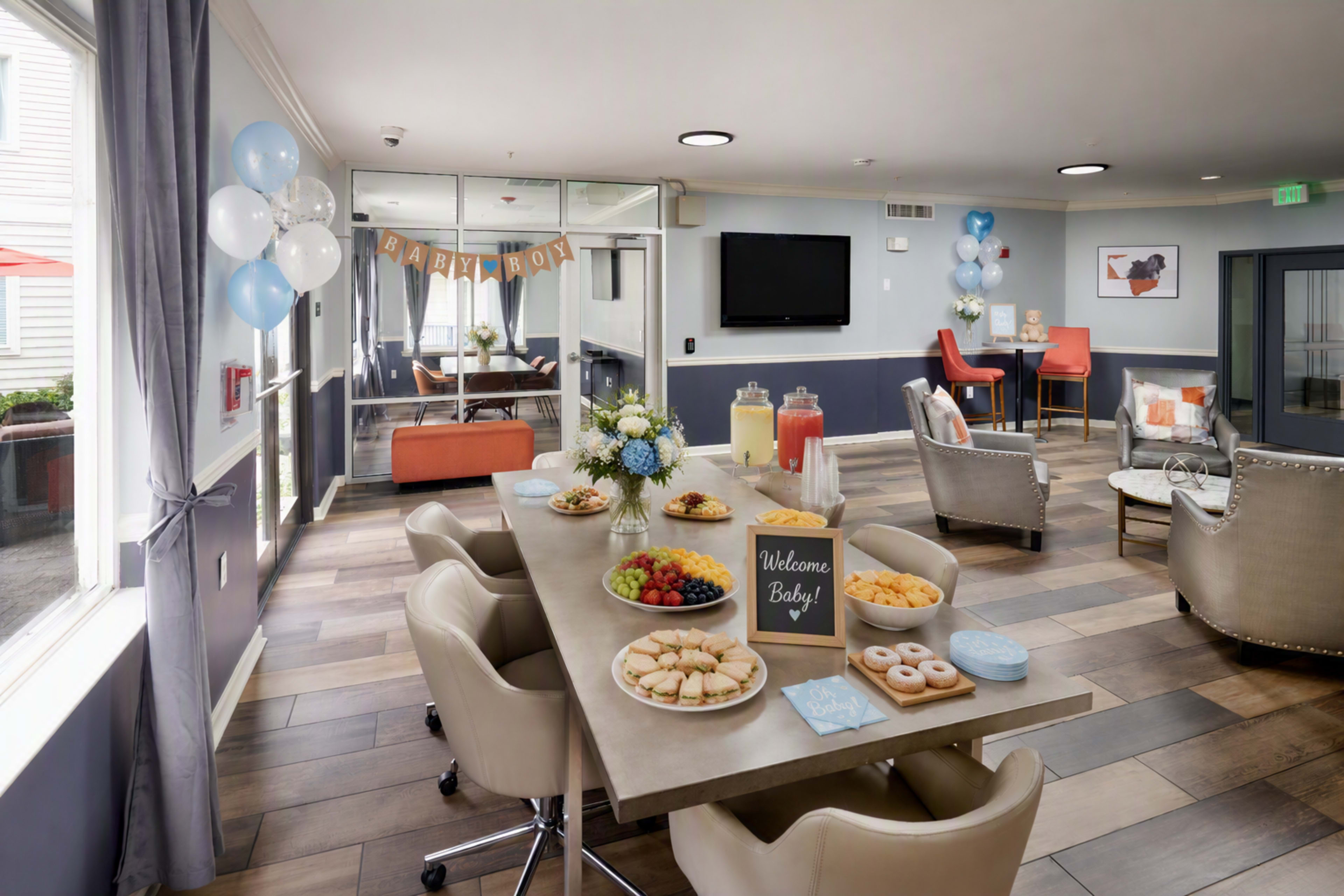 The image shows a spacious gathering area decorated for a baby shower, featuring a table with snacks, fruit, and drinks, surrounded by seating and balloons.