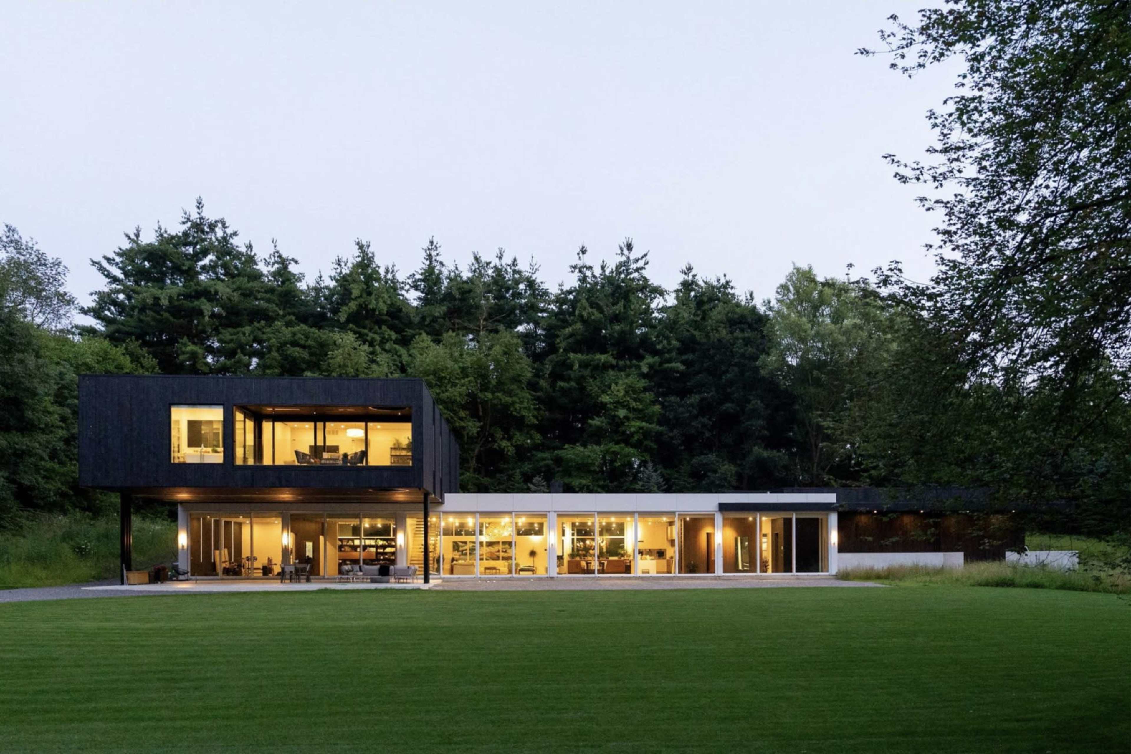 A modern black and white house with large windows is set against a backdrop of lush trees and a manicured lawn.