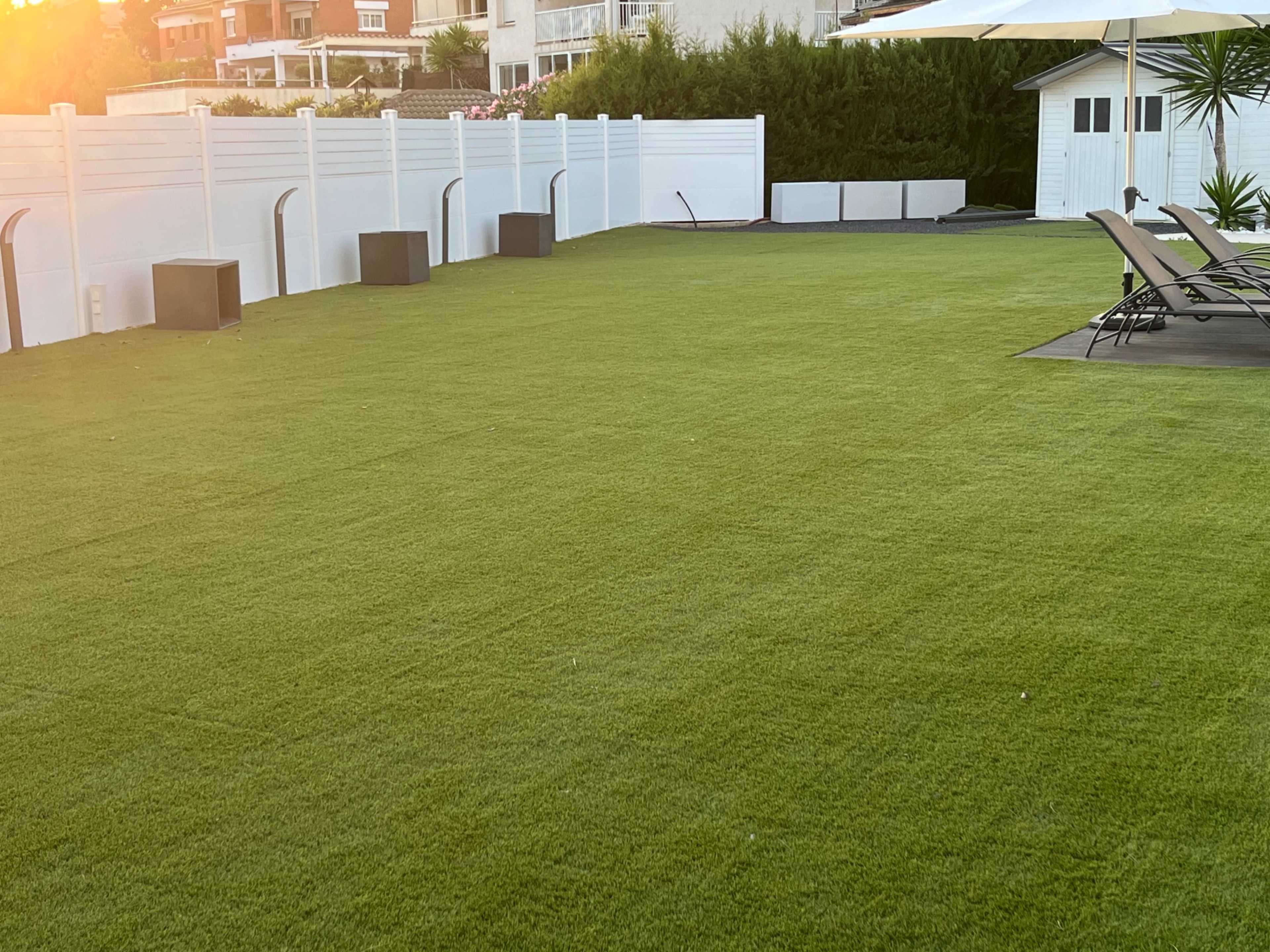 A green, artificial grass lawn bordered by a white fence, with lounge chairs and a small structure in the background.