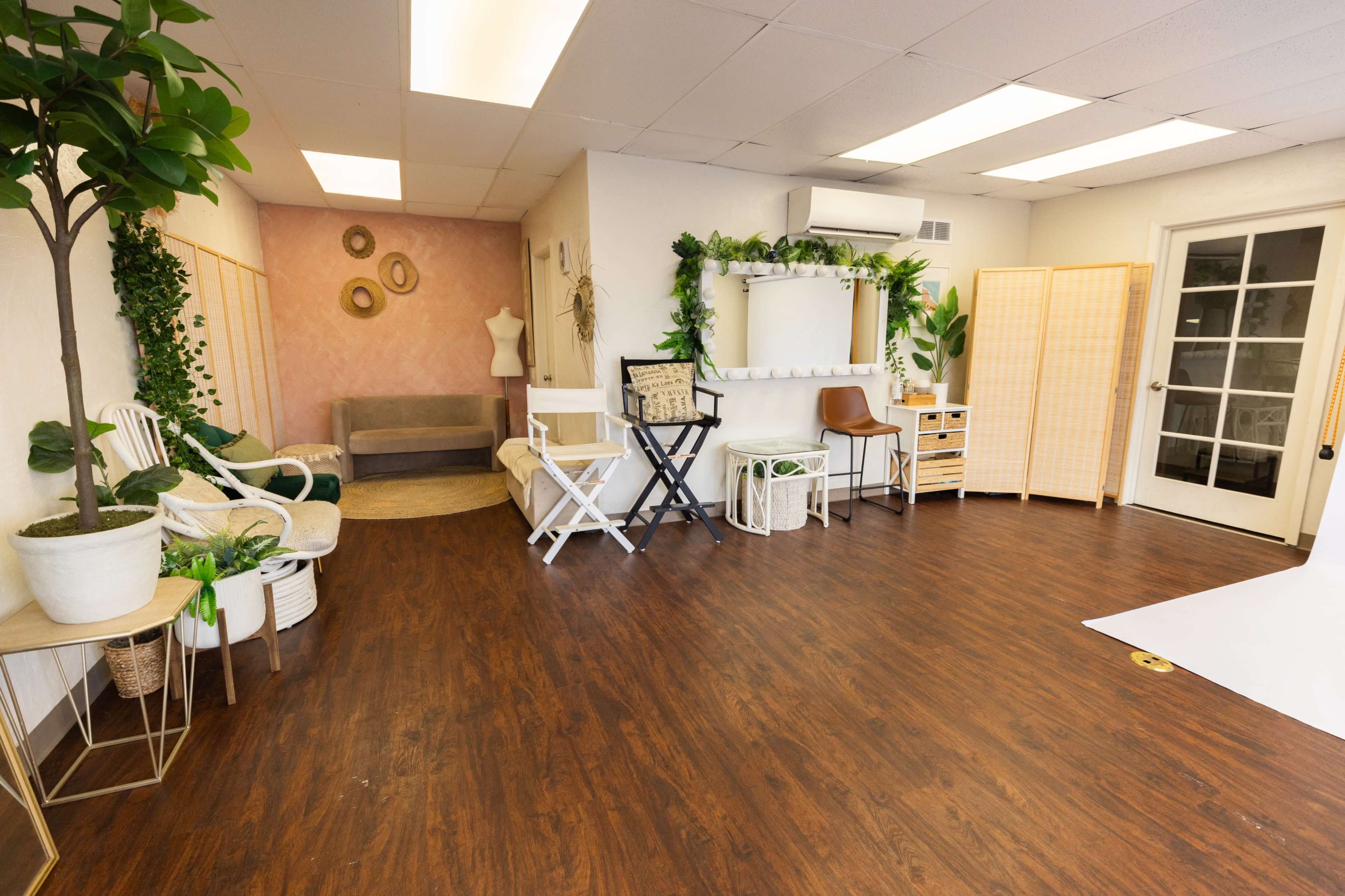 The image shows a spacious room with hardwood floors, featuring a seating area with chairs, a mirror, and decorative plants.