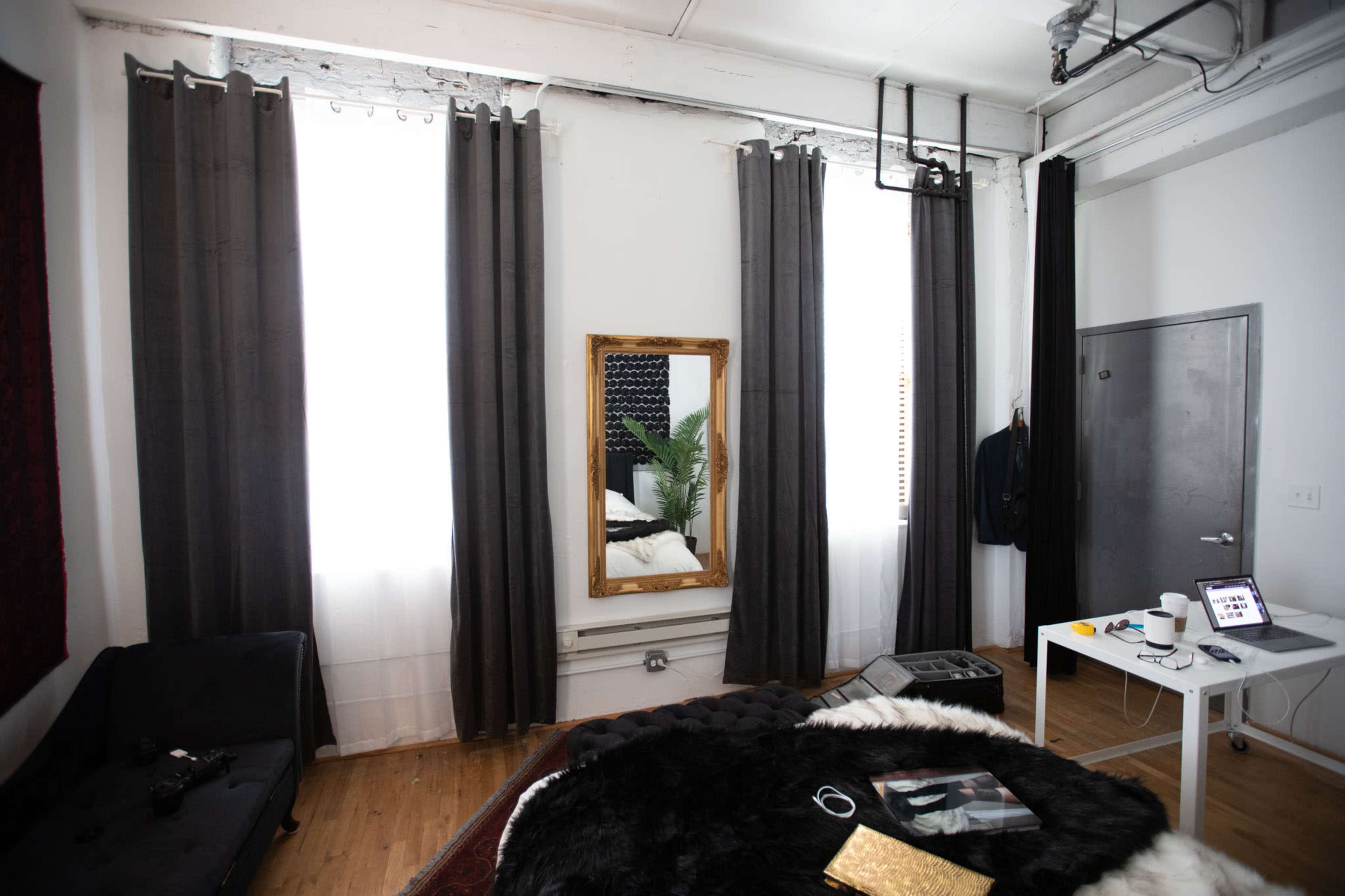 A room with dark curtains, a large mirror, a bed with a black and white blanket, and a small table with a laptop and some items on it.