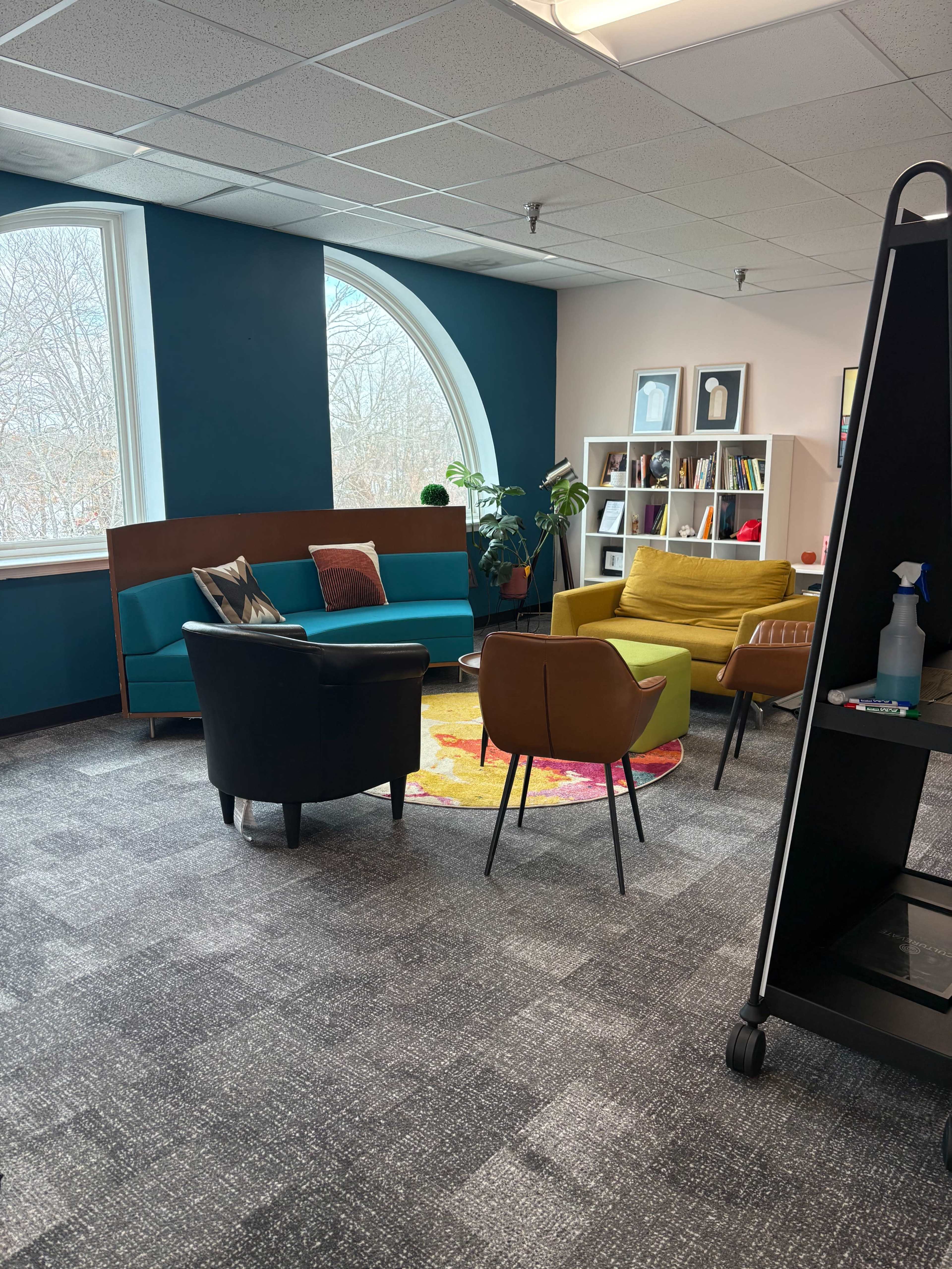 The image shows a modern office lounge area with colorful chairs, a bookshelf, and large windows allowing natural light to enter.