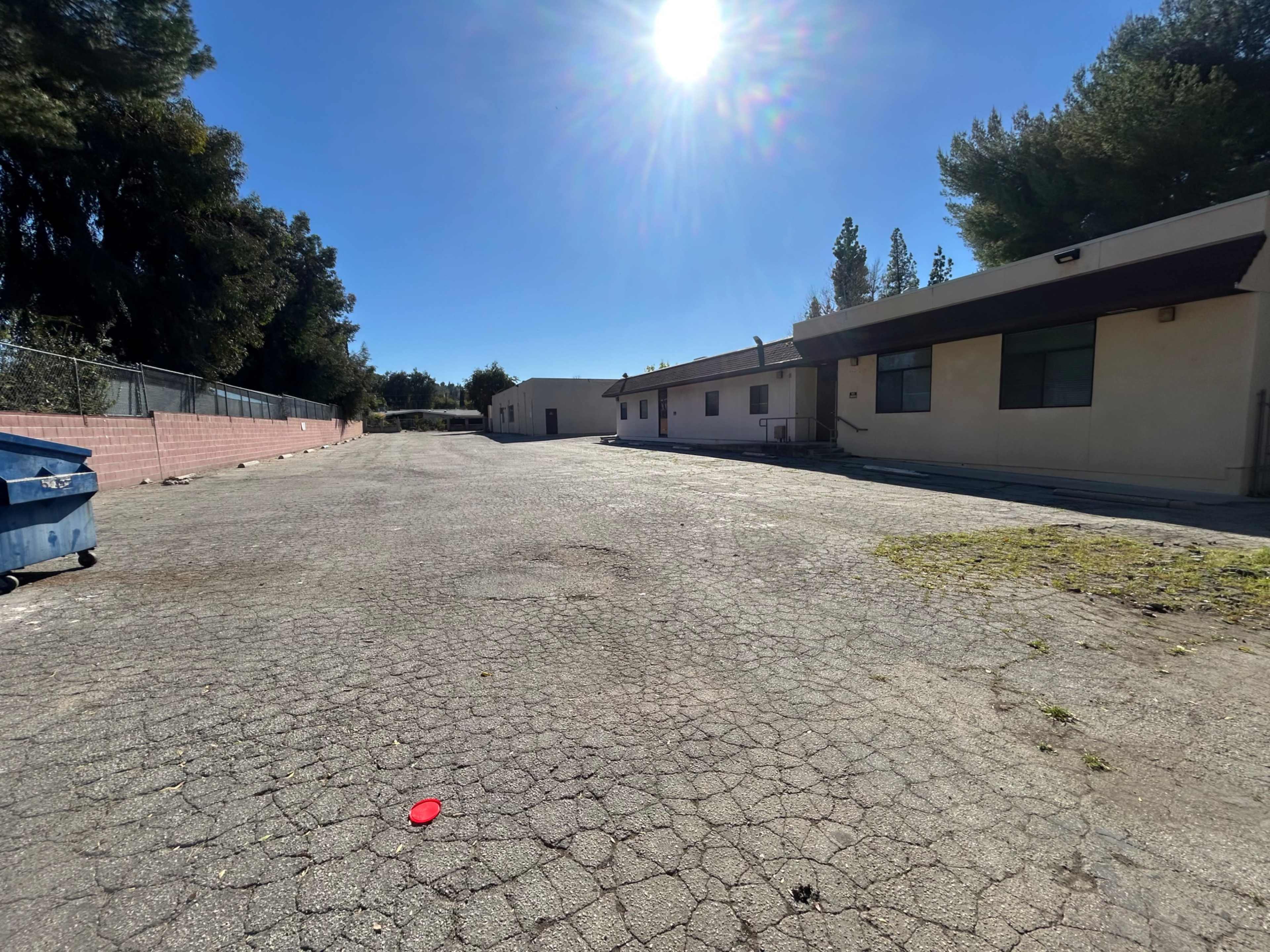 The image shows a wide, empty asphalt area with a few buildings on one side and a blue dumpster on the left.
