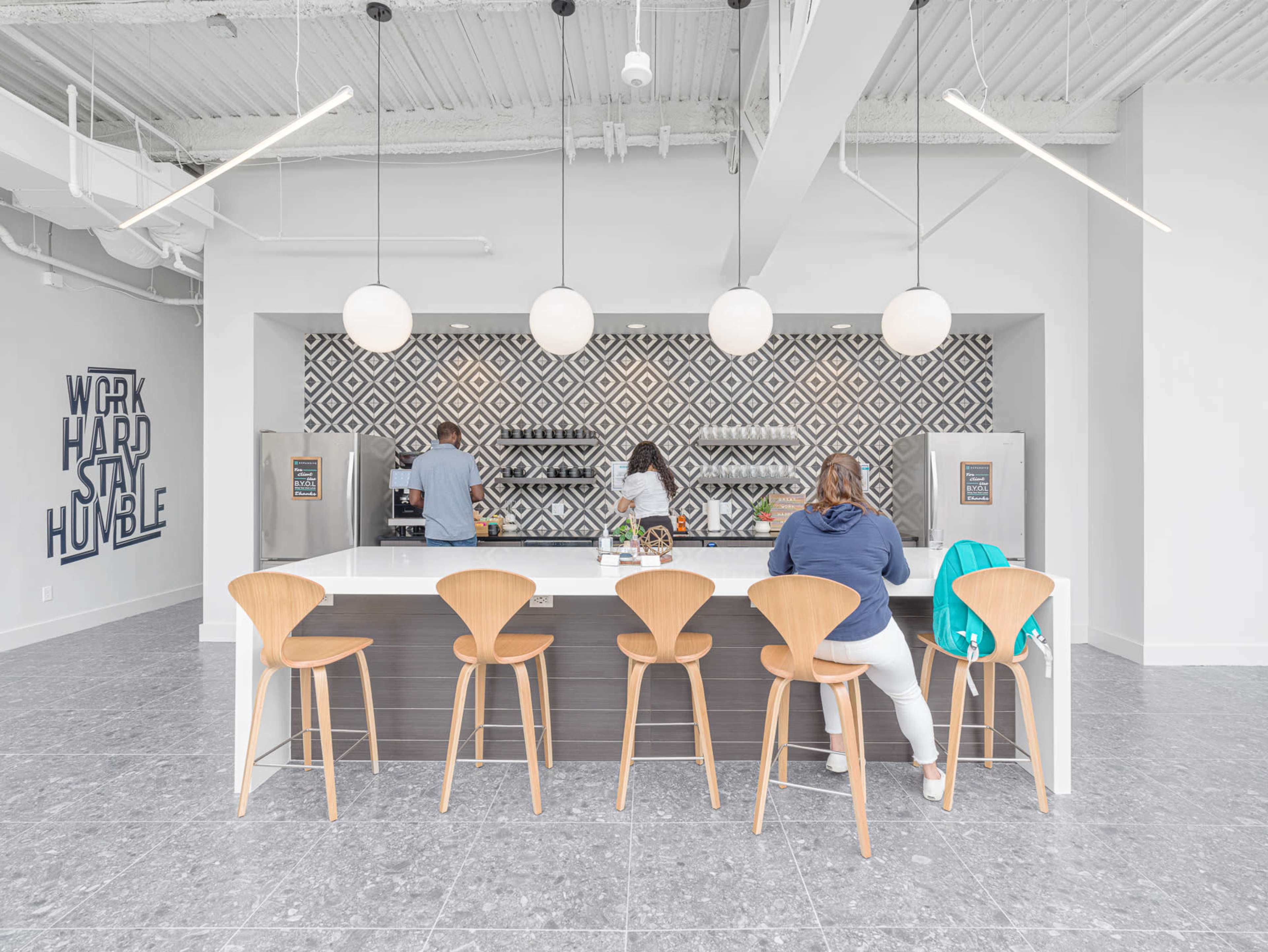 A modern kitchen area features a long island with wooden stools, two individuals are seated, and geometric tile decor lines the back wall.