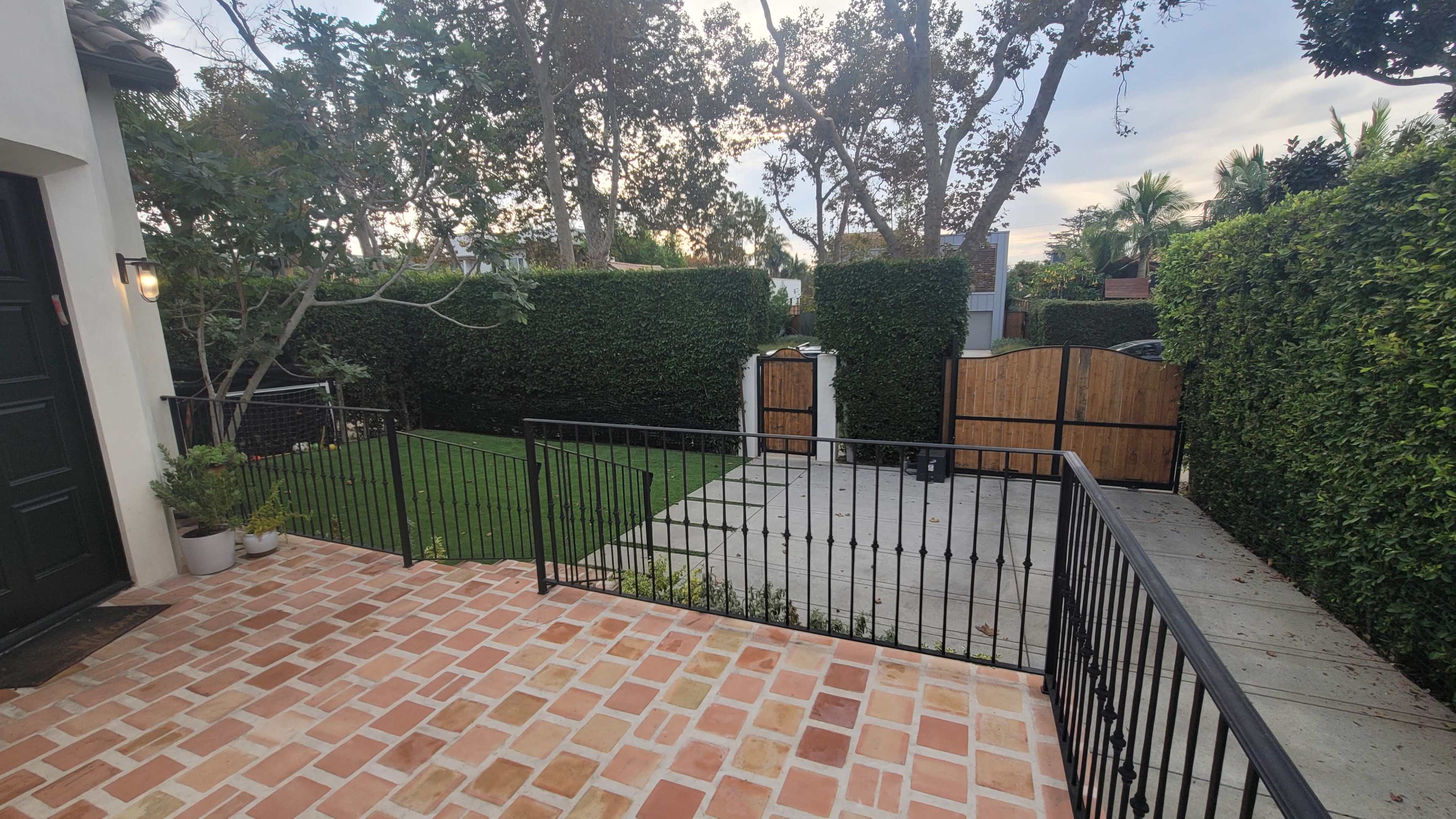 A brick-paved patio overlooks a neatly trimmed lawn and a gated area surrounded by tall hedges and trees.