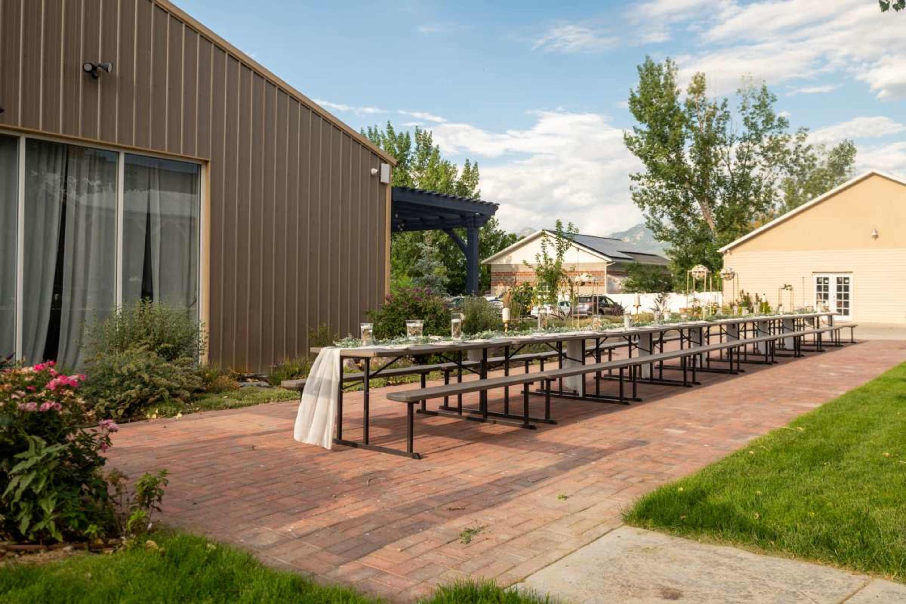 An outdoor dining setup features a long table lined with glasses and decorations, set against a backdrop of buildings and greenery.