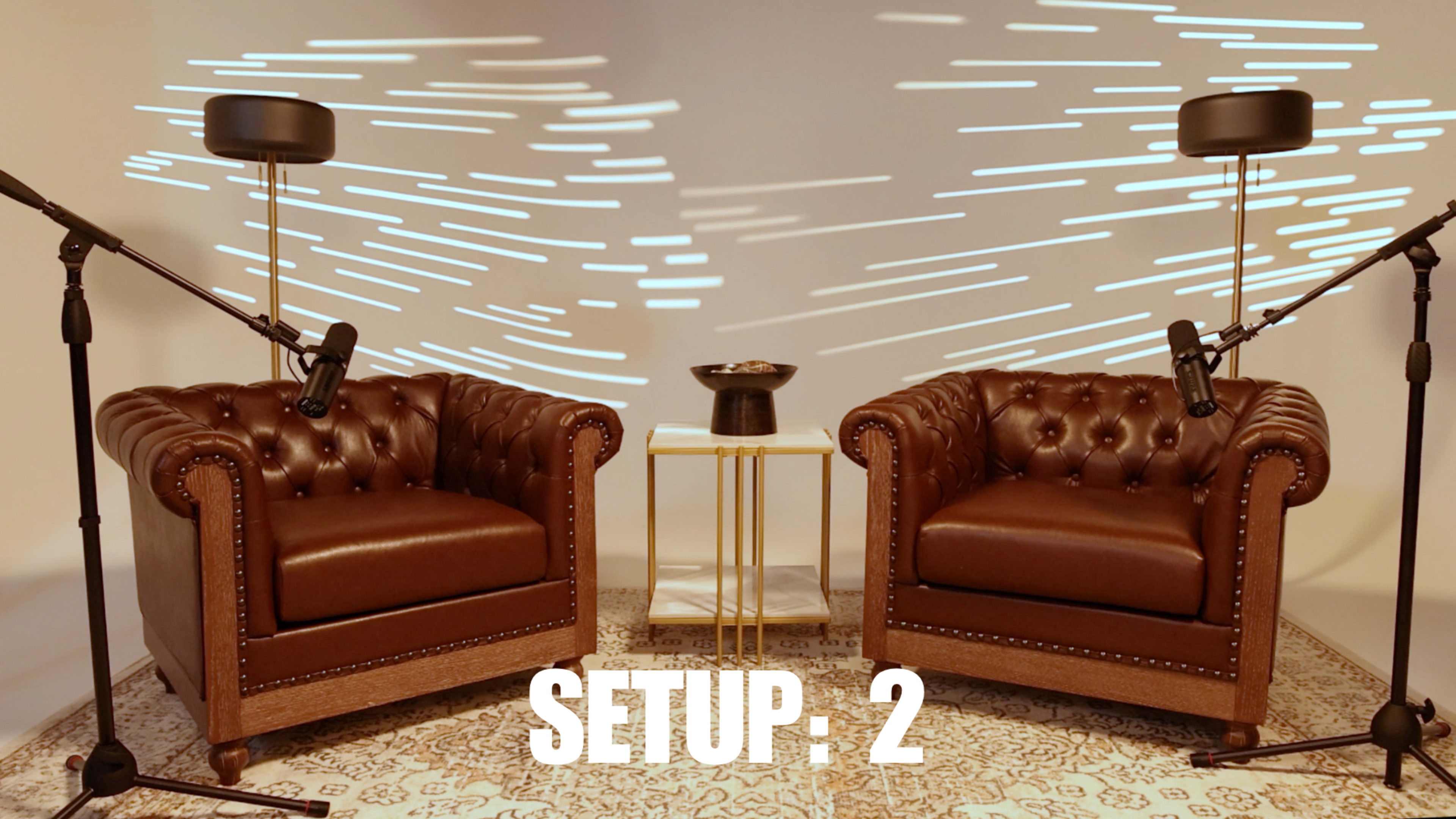The image shows two brown leather chesterfield armchairs facing each other, separated by a small table with a decorative bowl, and two microphones on stands positioned near each chair against a softly lit background that features linear patterns.
