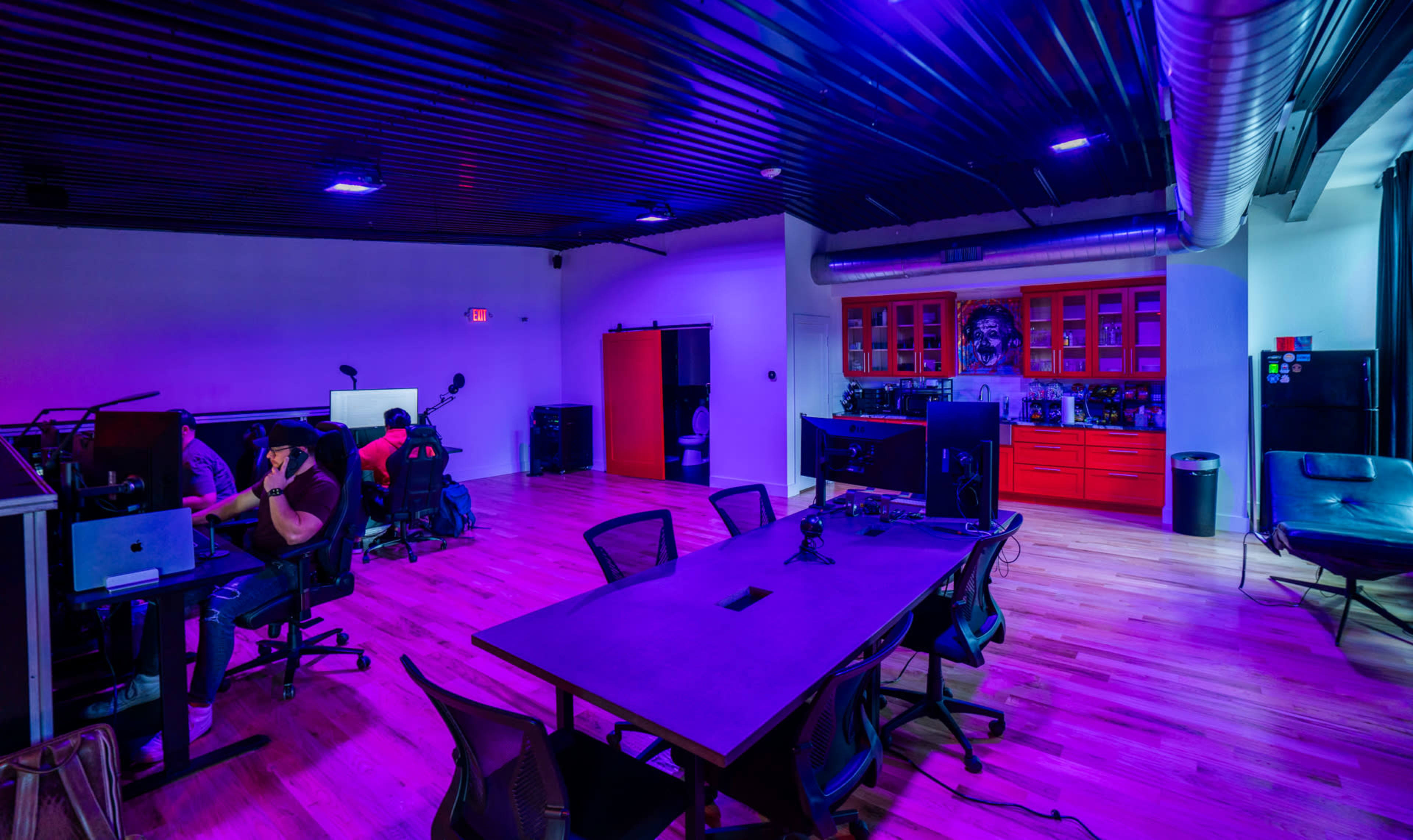 The image shows a modern office space with purple lighting, featuring several workstations, a large central table, and a red cabinet against the back wall.