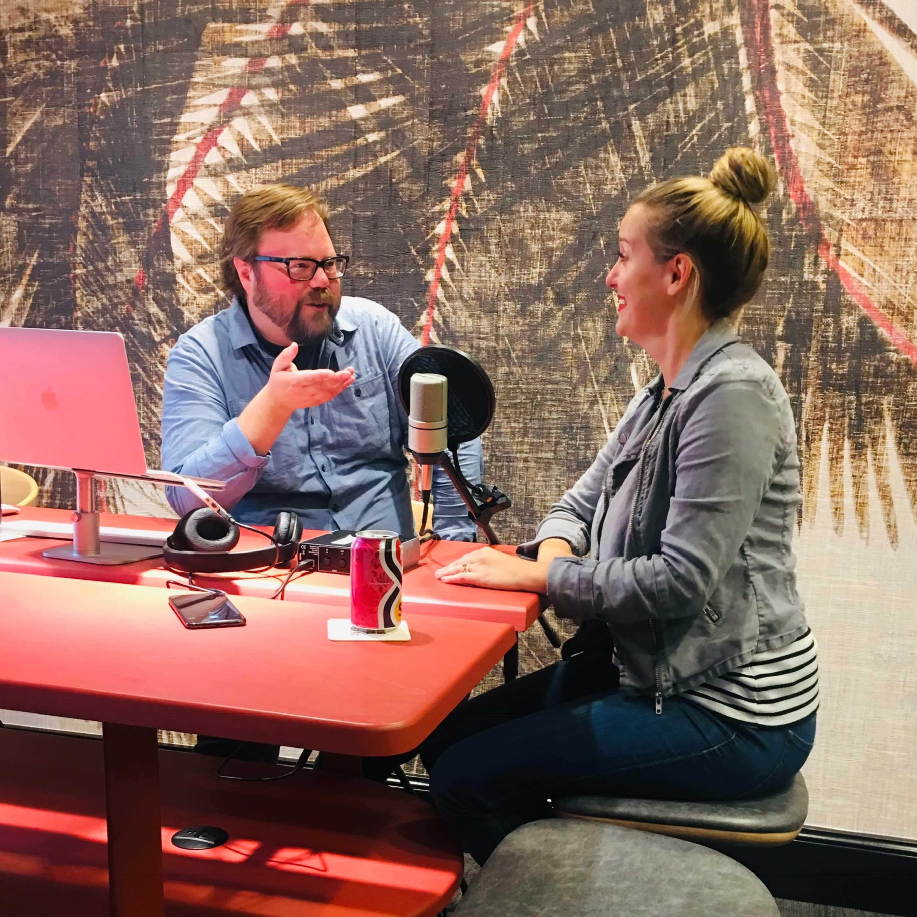 A man and a woman are sitting at a red table in a podcast studio, engaged in conversation with a microphone between them.