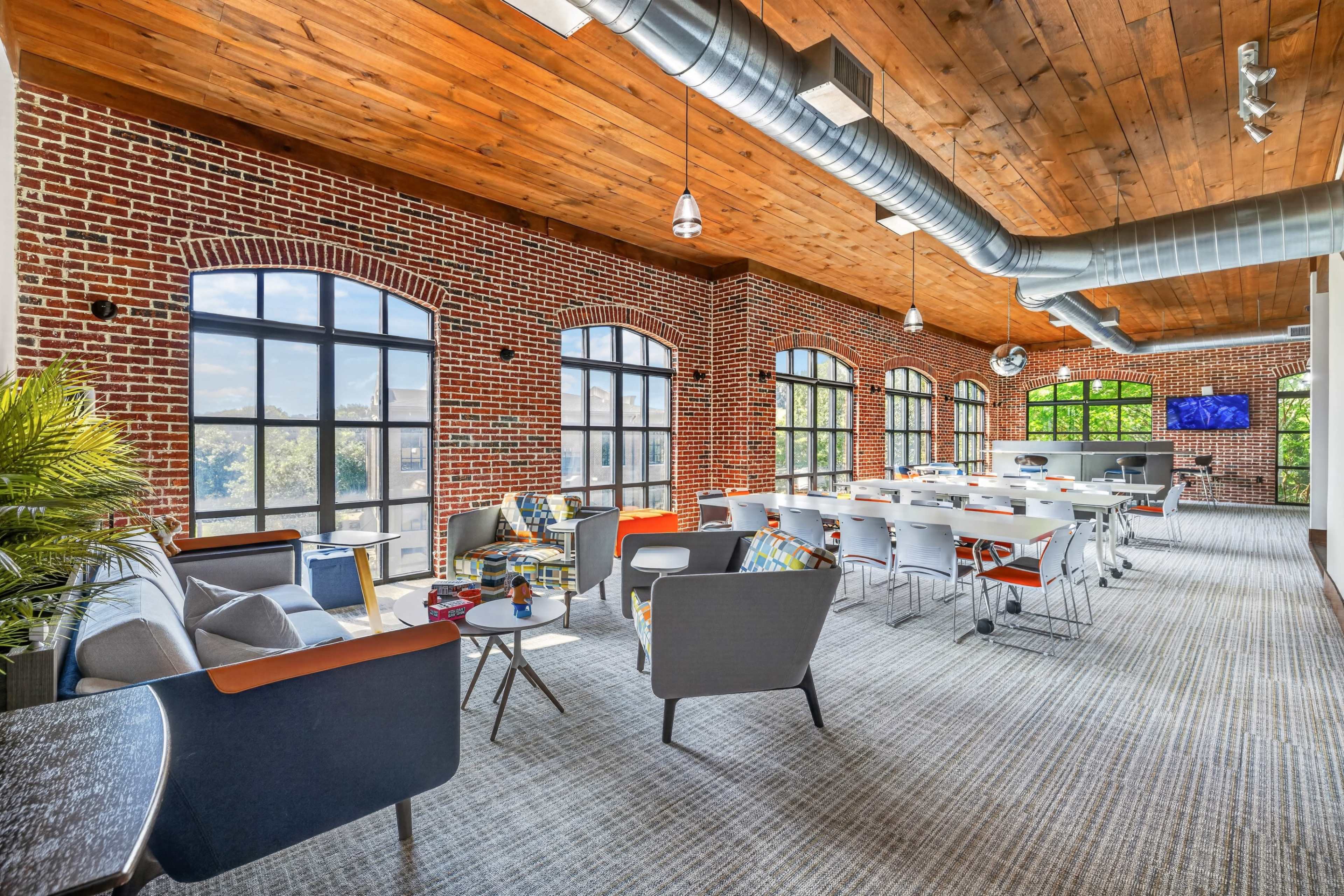 The image shows a modern office space with large windows, exposed brick walls, and a mix of seating arrangements including chairs and tables.