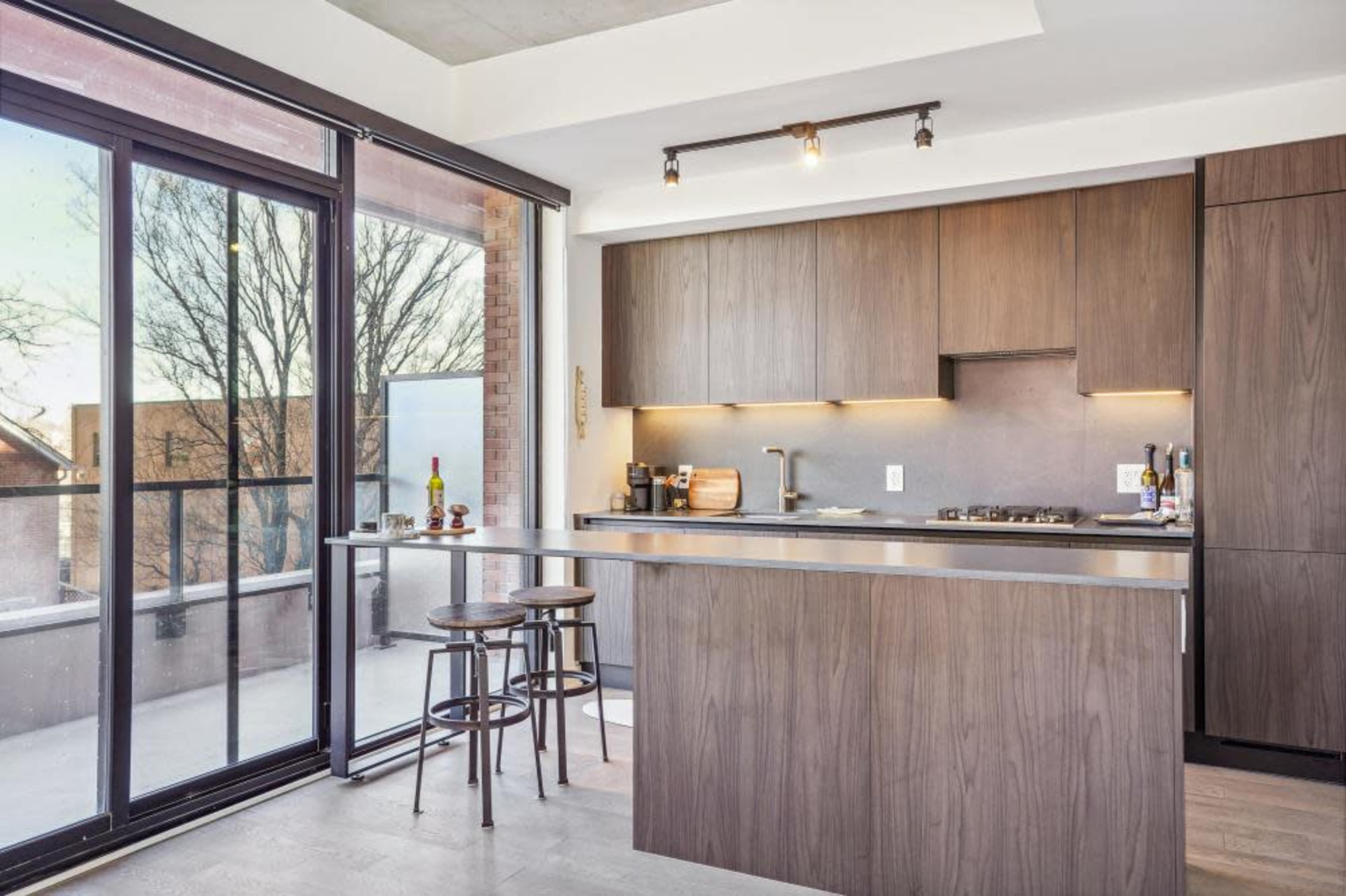 A modern kitchen features a wooden island, bar stools, and large glass doors opening to a balcony with trees visible outside.