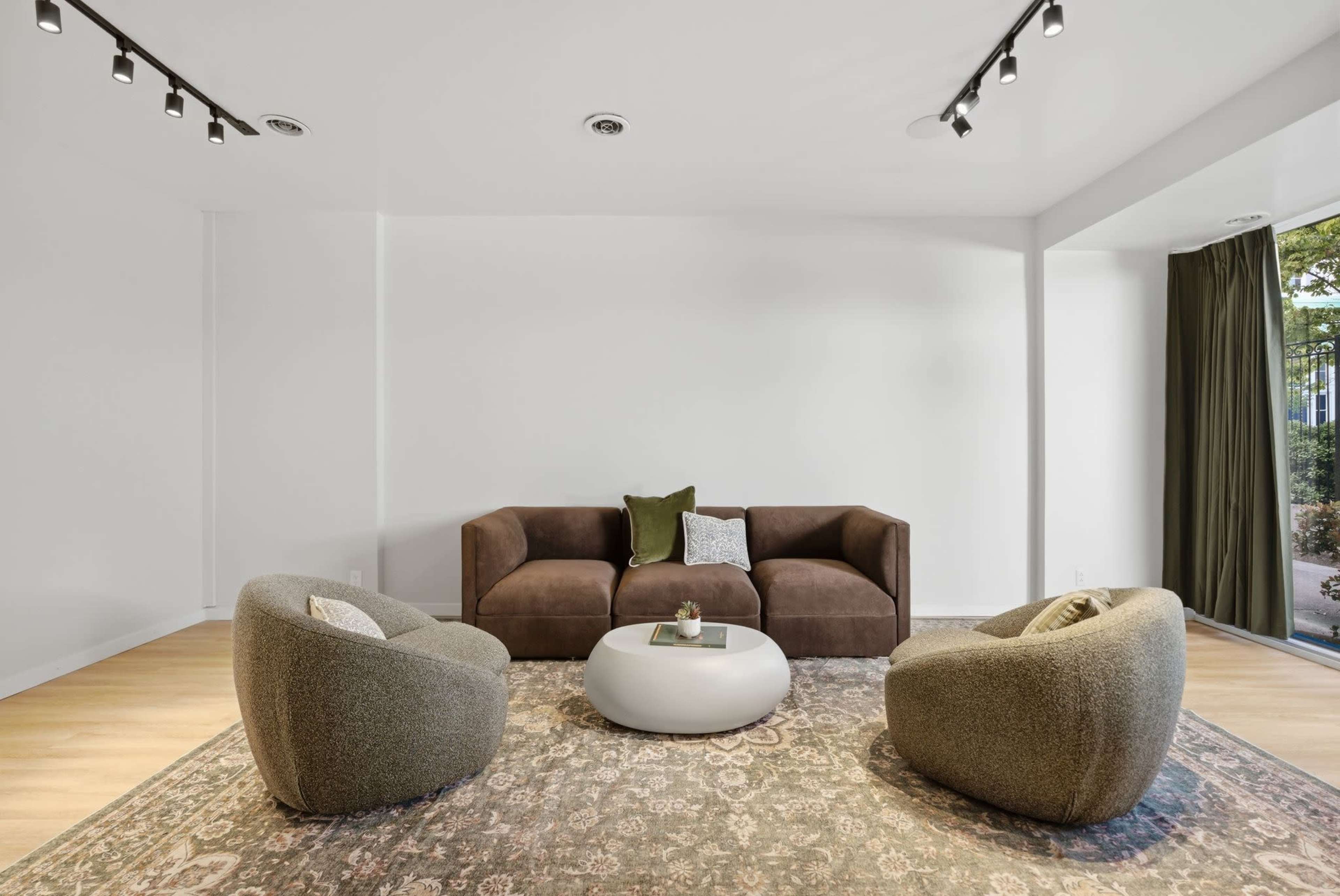 The image shows a modern living room featuring a brown sofa, two curved armchairs, a round coffee table, and a large area rug, with natural light coming through a window.