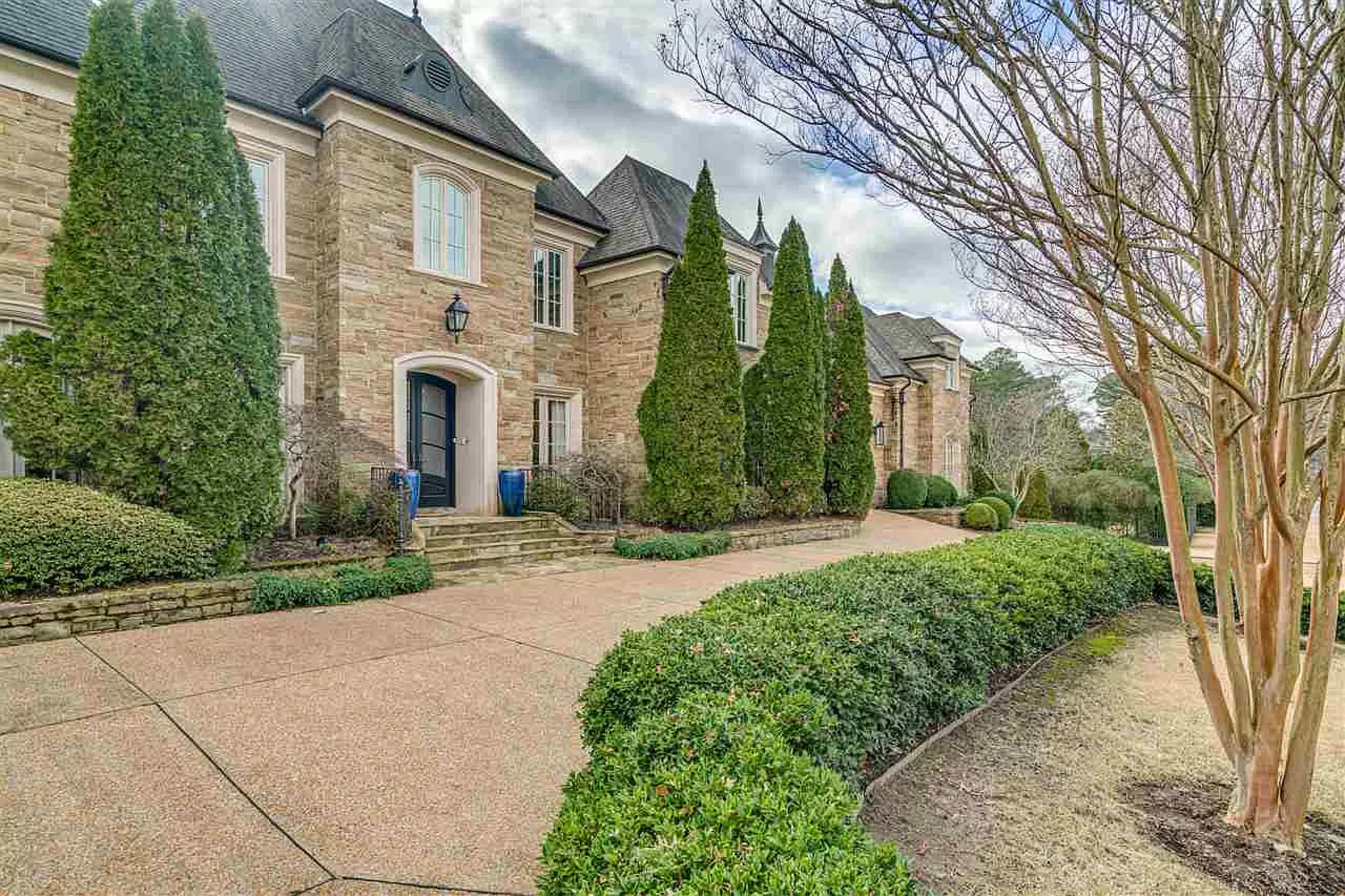 A large stone mansion with neatly trimmed shrubs and tall trees lining the driveway.