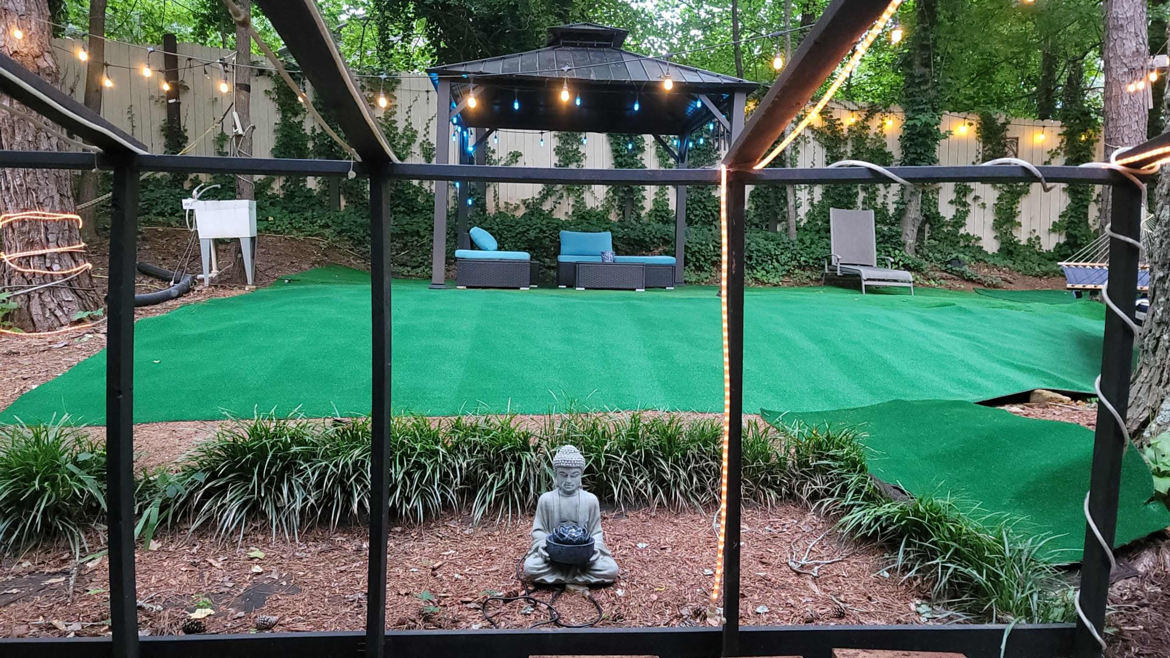A backyard features an artificial turf area with a gazebo, seating arrangement, and a statue of Buddha surrounded by greenery.