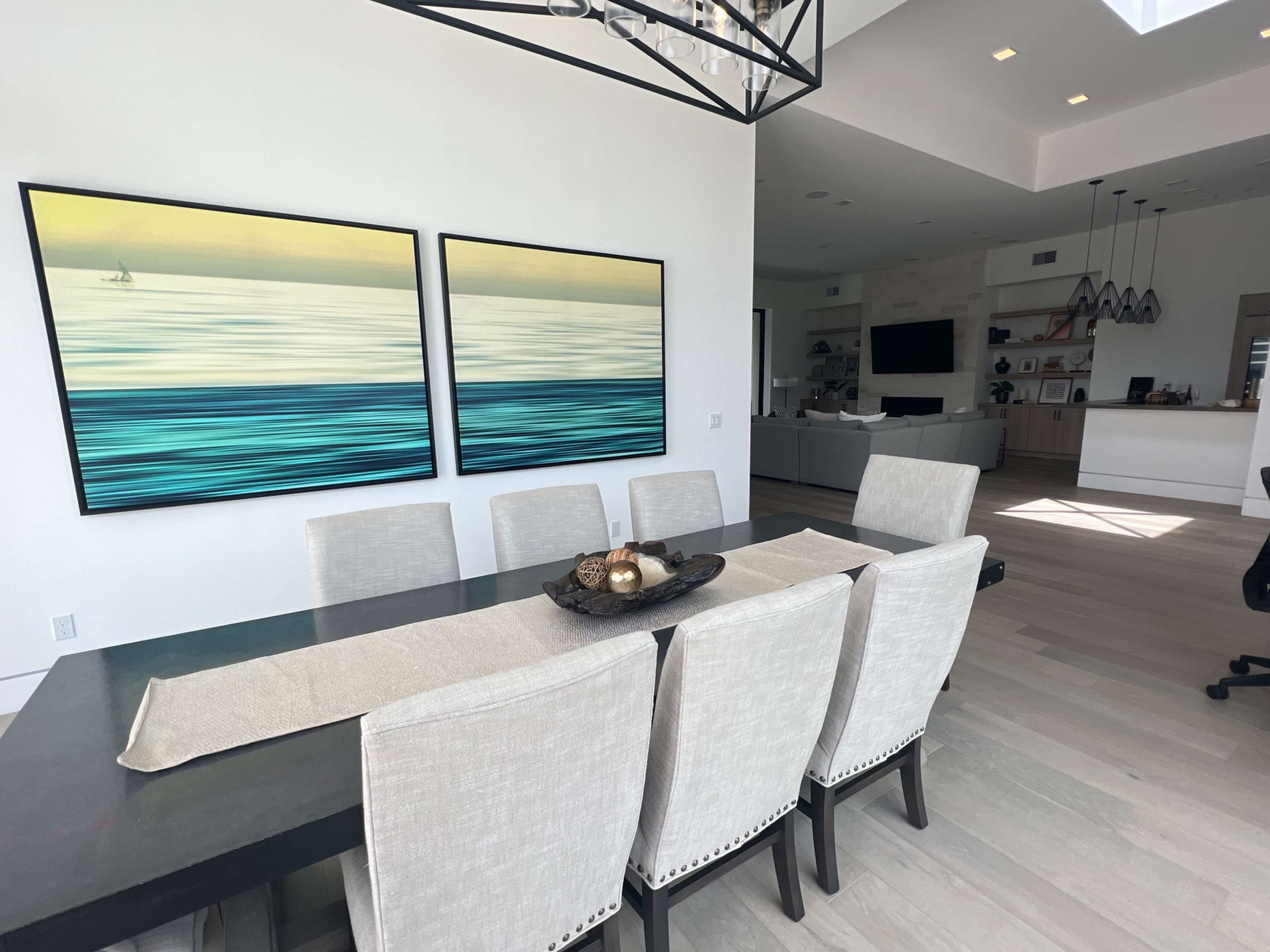 A modern dining area with a large table surrounded by light-colored chairs, featuring two abstract paintings on the wall and a view into a living space with a sofa and kitchen in the background.