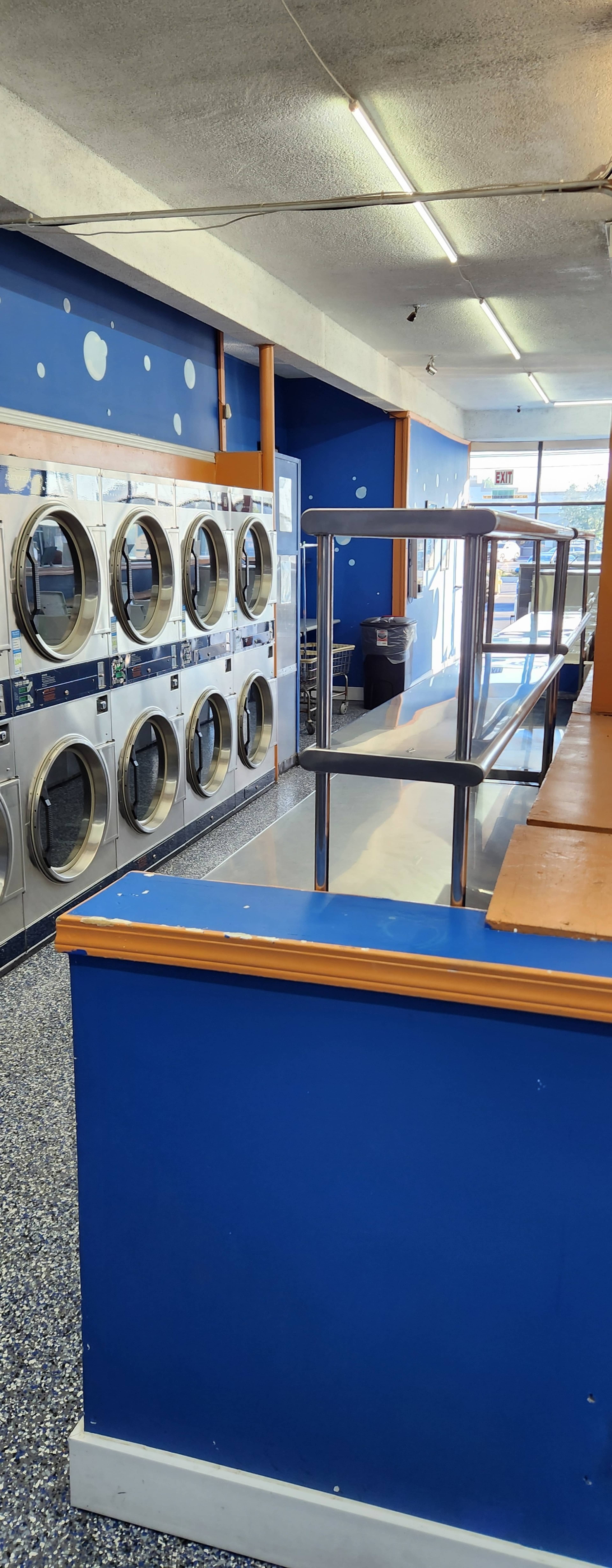 The image shows the interior of a laundromat with rows of washing machines along a blue wall and a counter in the foreground.
