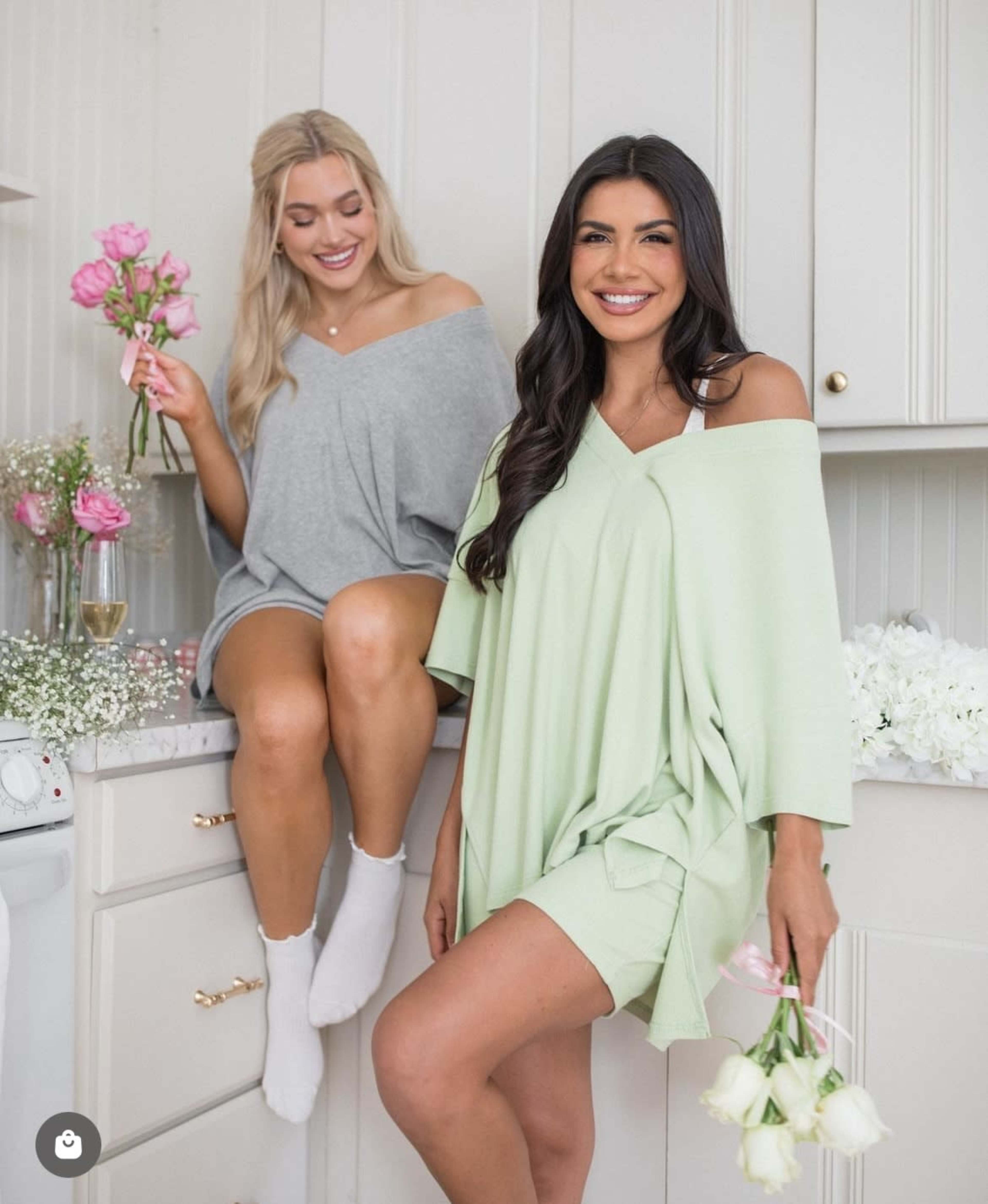 Two women pose in a kitchen, one sitting on a countertop holding flowers and the other standing with a bouquet.