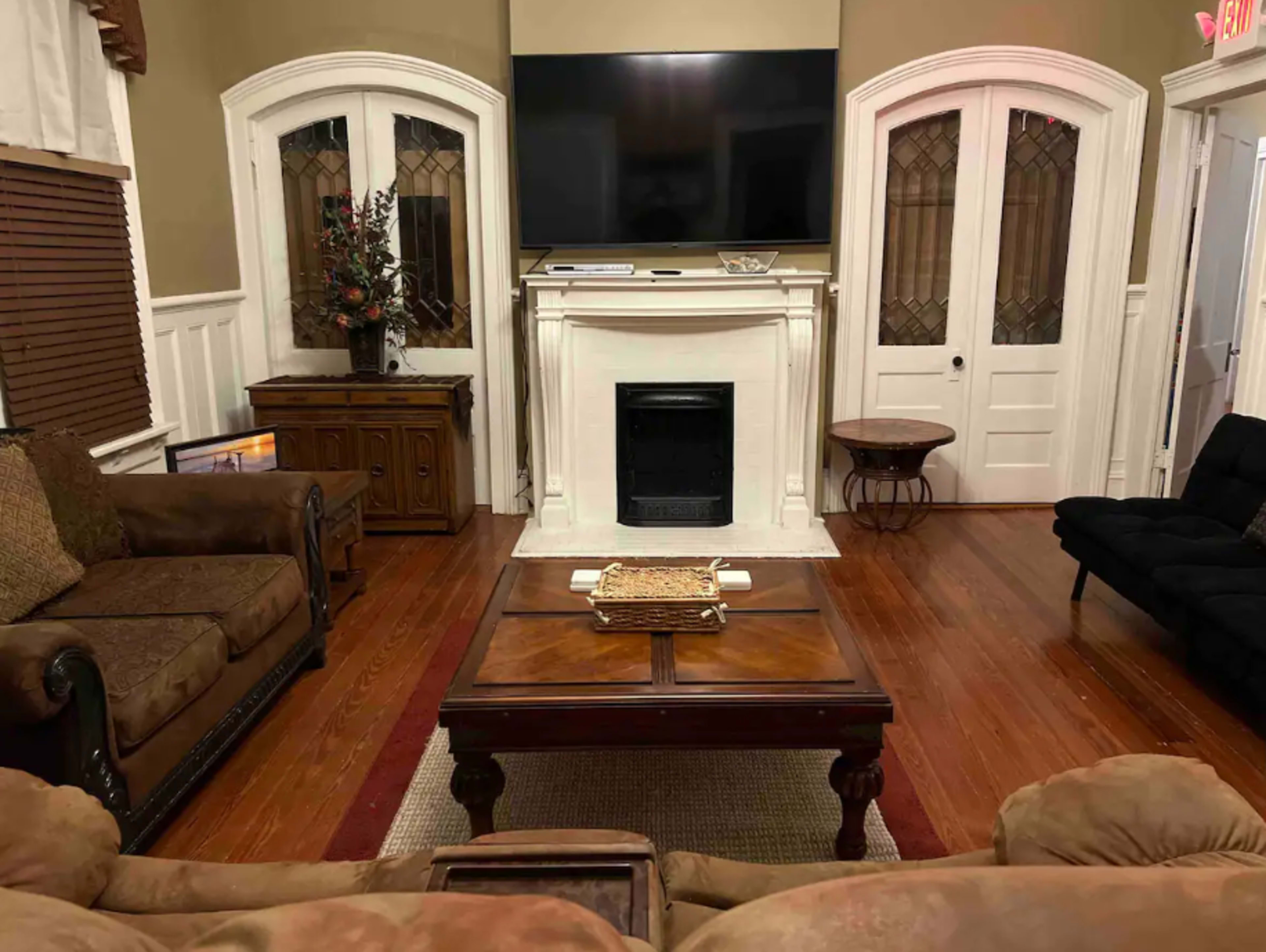 The living room features a fireplace beneath a large mounted television, flanked by two identical doors, with a coffee table at the center surrounded by couches.