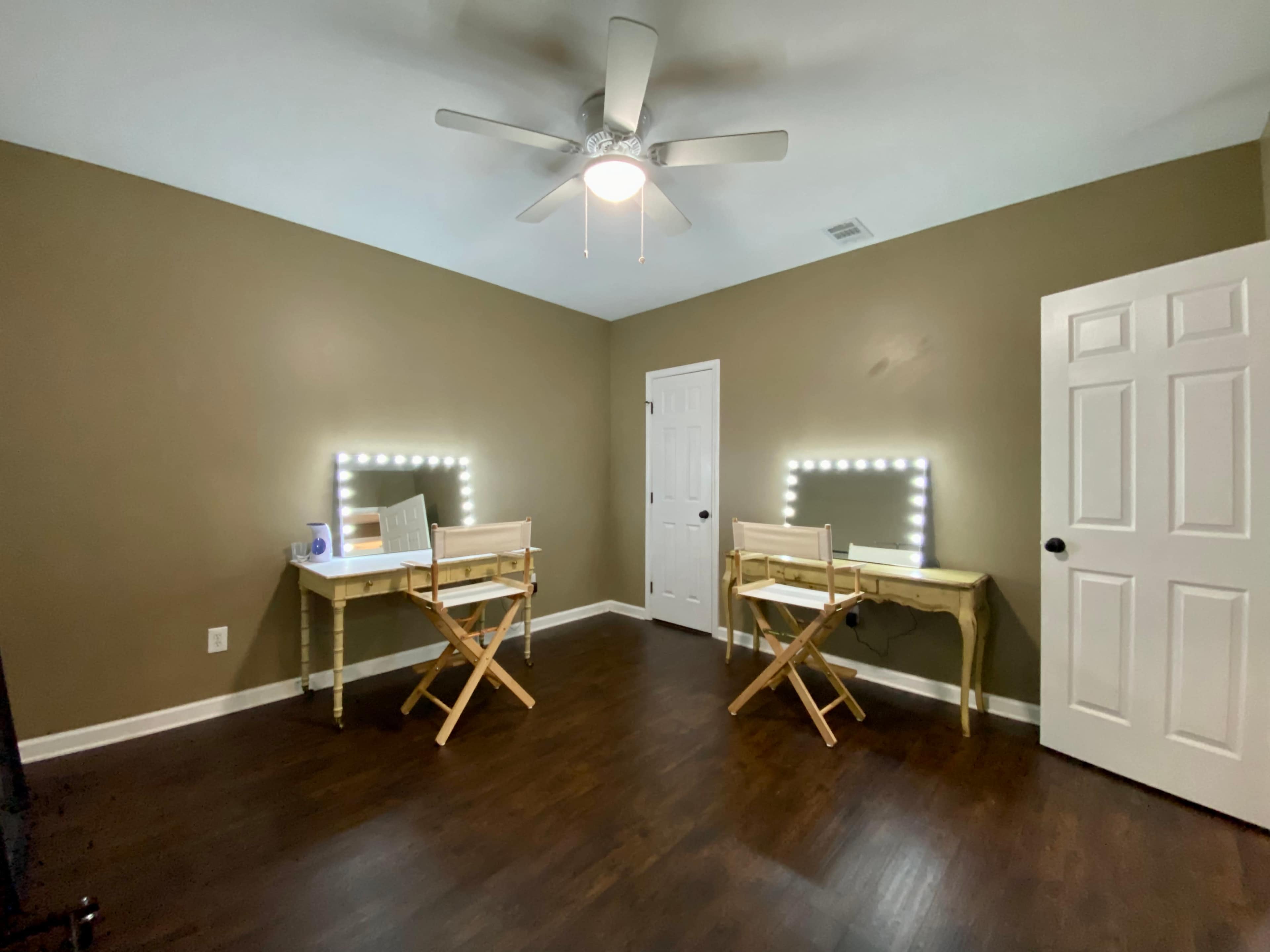 The room features two wooden folding tables with illuminated mirrors and a ceiling fan, set against a brown-painted wall.